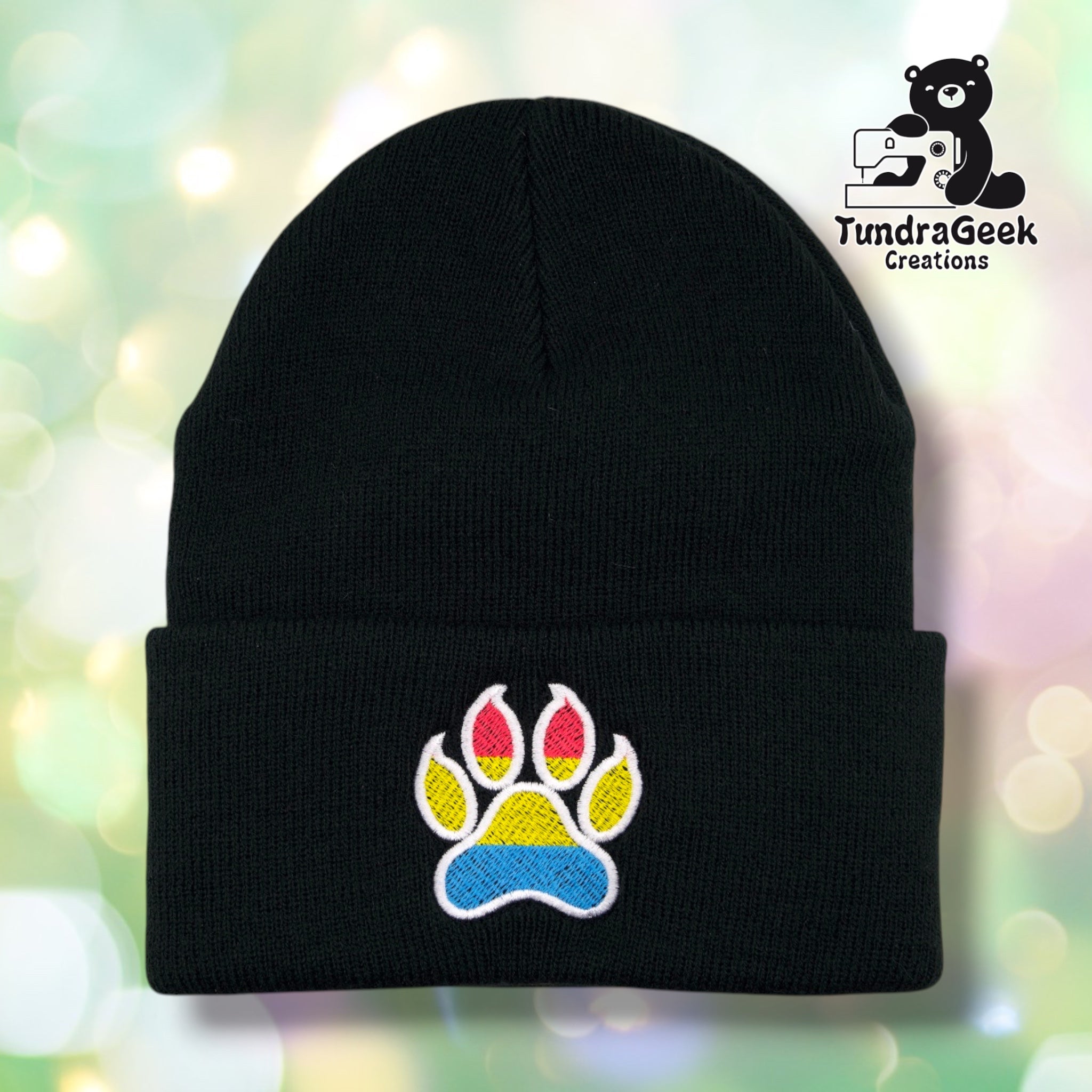 A black knit beanie with a folded cuff featuring an embroidered paw print in yellow, red, and blue with white outlines. The beanie is centered against a soft, blurred green bokeh background with a 'TundraGeek Creations' logo in the top right corner.