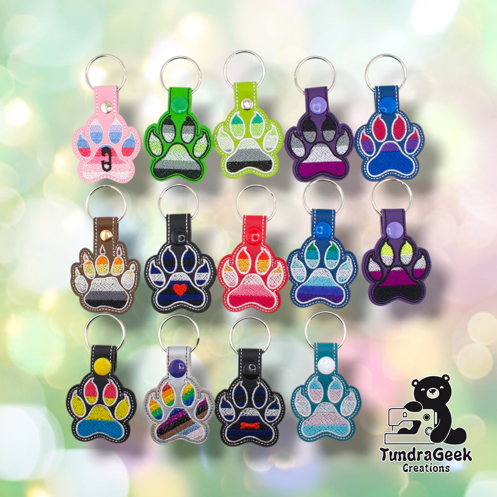 Fourteen embroidered vinyl paw-shaped pride keychains arranged in a three-row grid on a soft bokeh background. The collection includes a wide range of flag colorways. Row one: ABDL (pink), Agender canine-style (dark green), Aromantic (lime green), Asexual (purple), Bisexual (blue). Row two: Bear (brown), Leather (black with heart), Lesbian (red), MLM (blue), Non-Binary (purple). Row three: Pansexual (black), Progress Pride (silver), Pup (black with bone), Transgender (teal). All keychains have silver split 