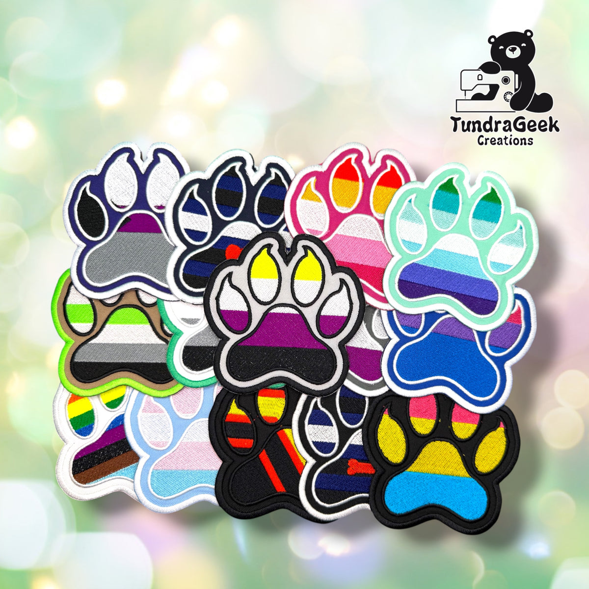 A collection of approximately fifteen embroidered paw-shaped pride patches spread out on a soft bokeh background. The patches display a wide variety of pride flag color combinations including rainbow, bisexual, pansexual, non-binary, asexual, leather, agender, progress pride, and others. Each patch features bold embroidered flag stripes within a paw print shape with a contrasting outline.
