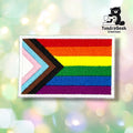 Front view of a rectangular patch with a white merrowed edge, embroidered with the Progress Pride flag — a chevron of white, pink, light blue, brown, and black stripes pointing right into a six-color rainbow field (red, orange, yellow, green, blue, purple).