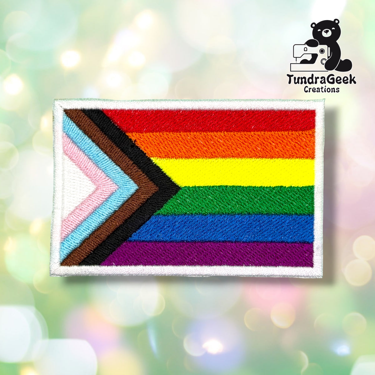 Front view of a rectangular patch with a white merrowed edge, embroidered with the Progress Pride flag — a chevron of white, pink, light blue, brown, and black stripes pointing right into a six-color rainbow field (red, orange, yellow, green, blue, purple).