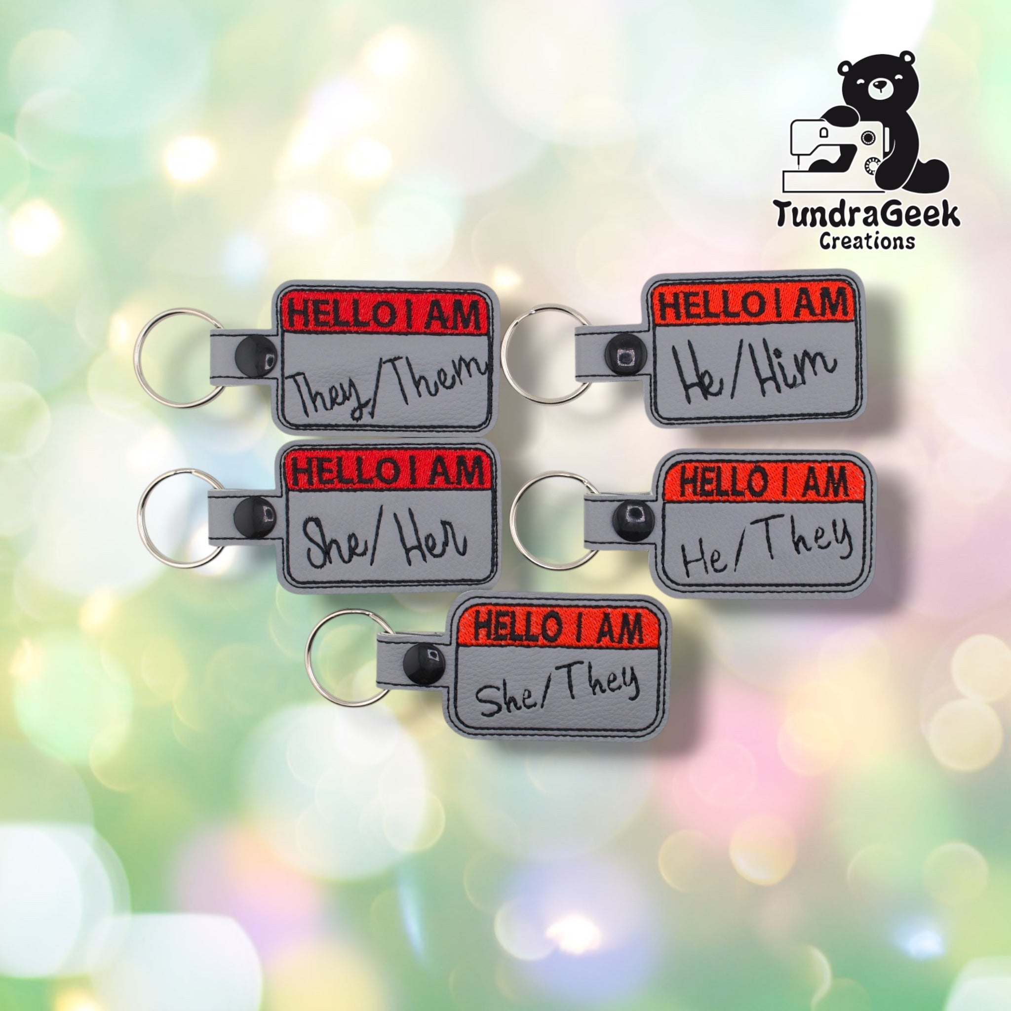 Five nametag-style keychains grouped together, each reading "HELLO I AM" in red with a different pronoun set embroidered below in black — They/Them, He/Him, She/Her, He/They, and She/They.