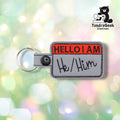 Front view of a gray nametag-style keychain with "HELLO I AM" embroidered in red at the top and "He/Him" in black hand-lettered-style text below, finished with a black plastic snap closure and split ring.