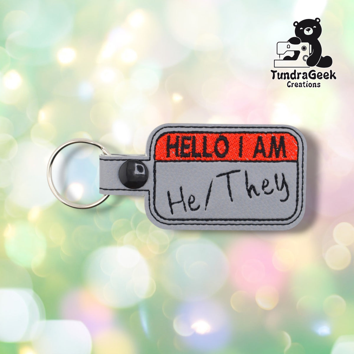 Front view of the same nametag-style keychain design with "He/They" embroidered in black beneath the red "HELLO I AM" header.