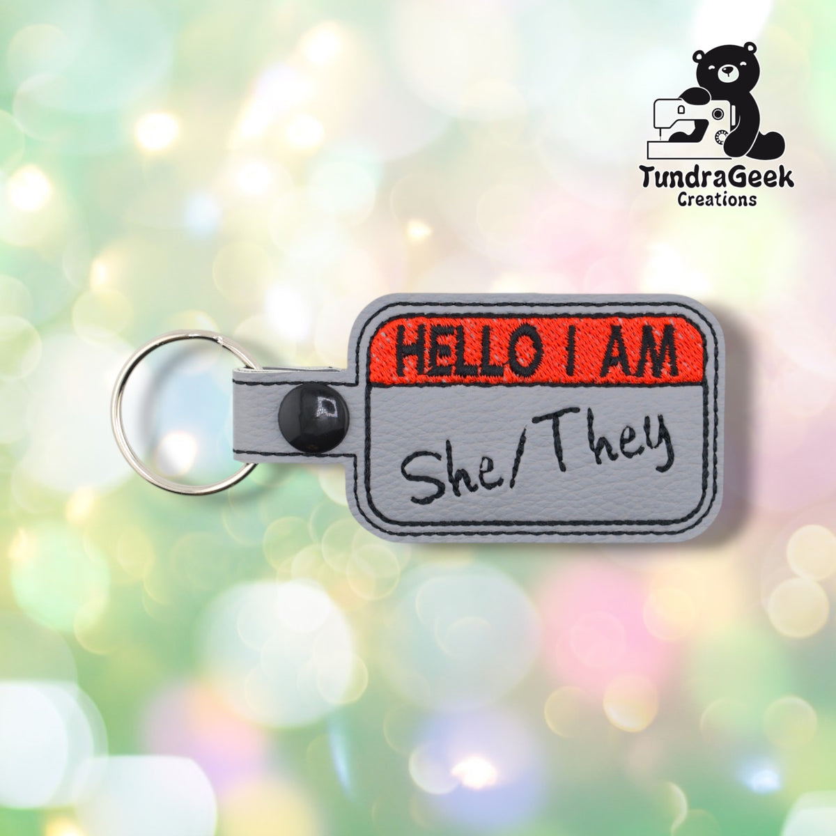 Front view with "She/They" embroidered in black beneath the red "HELLO I AM" header.