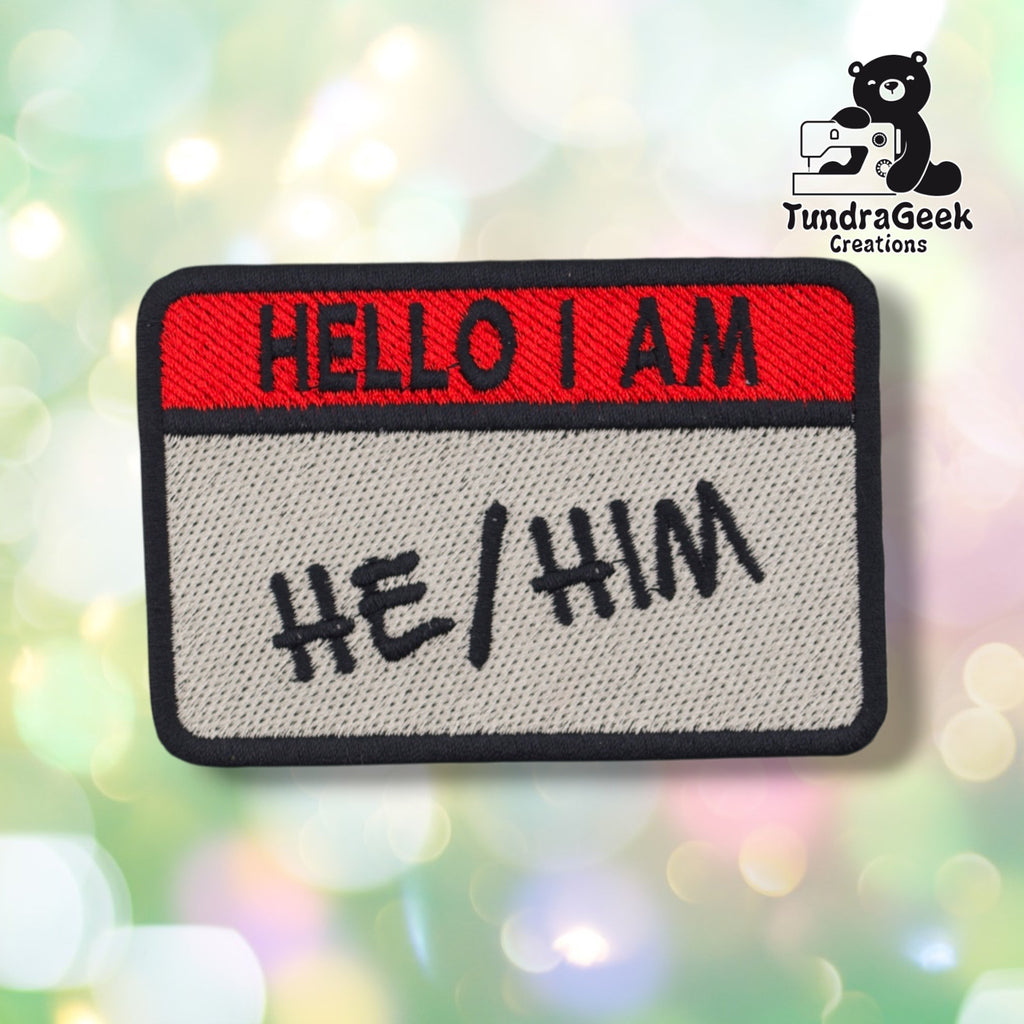 A rectangular embroidered patch with a red top band reading 'HELLO I AM' and a light gray lower section reading 'HE/HIM' in black script, shown on a blurred pastel bokeh background. A TundraGeek Creations logo appears in the top right corner.