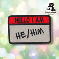 A rectangular embroidered patch with a red top band reading 'HELLO I AM' and a light gray lower section reading 'HE/HIM' in black script, shown on a blurred pastel bokeh background. A TundraGeek Creations logo appears in the top right corner.