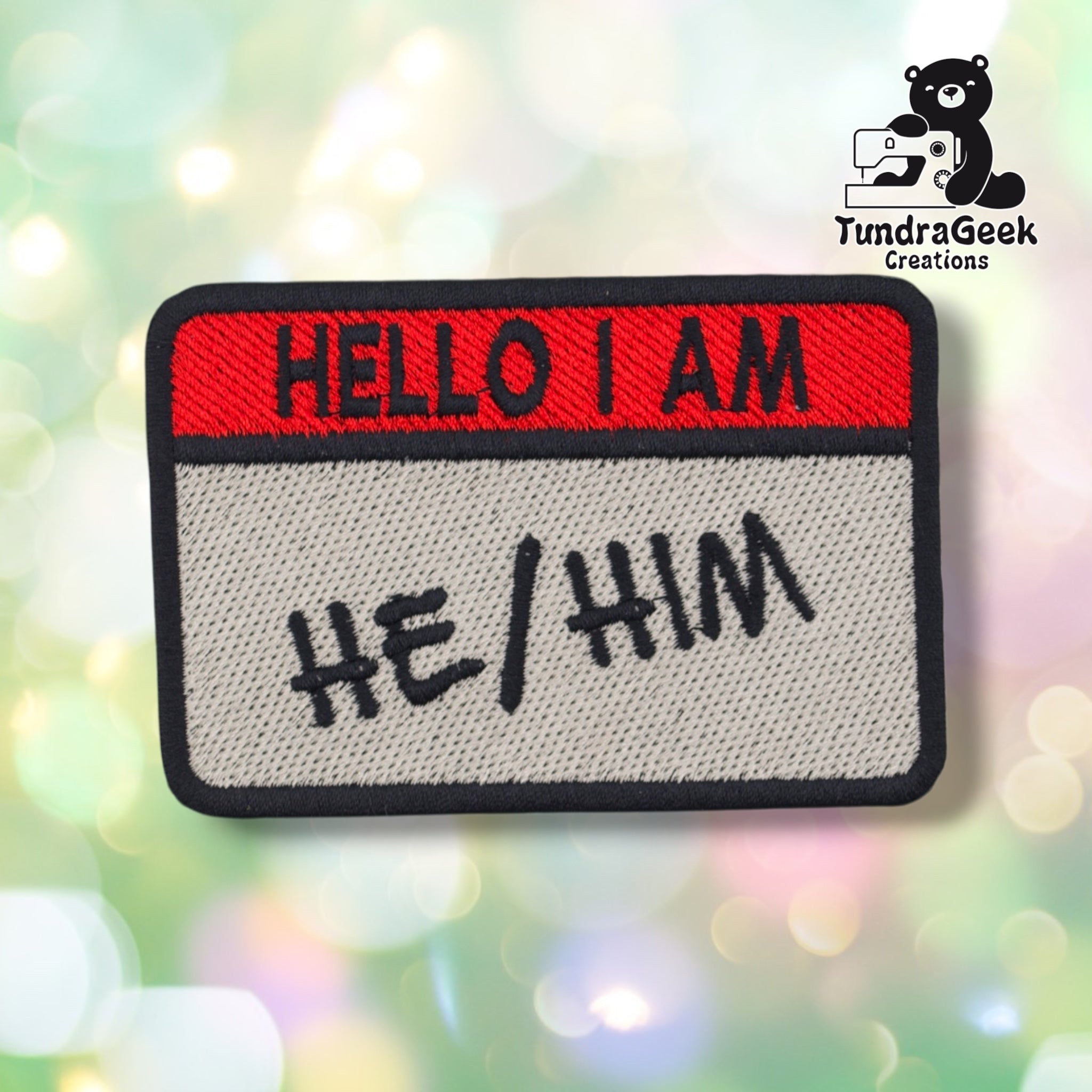 A rectangular embroidered patch with a red top band reading 'HELLO I AM' and a light gray lower section reading 'HE/HIM' in black script, shown on a blurred pastel bokeh background. A TundraGeek Creations logo appears in the top right corner.