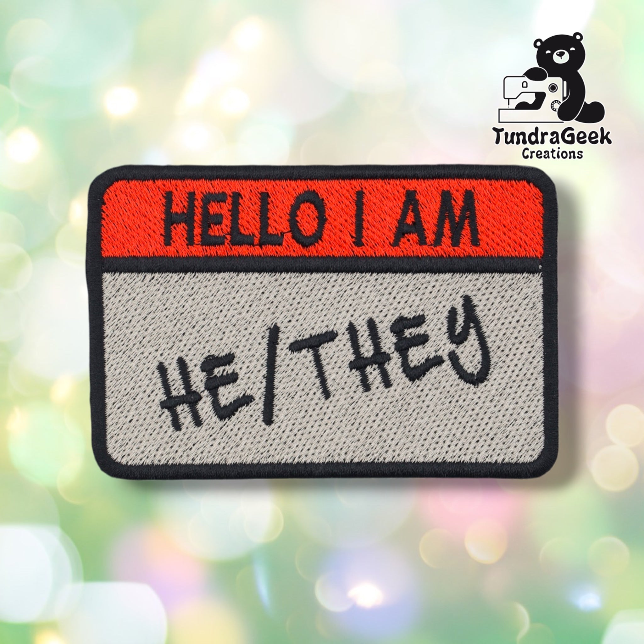 A rectangular embroidered patch with a red top band reading 'HELLO I AM' and a light gray lower section reading 'HE/THEY' in black script. The patch is centered on a soft multicolor bokeh background, with the TundraGeek Creations logo in the top-right corner.