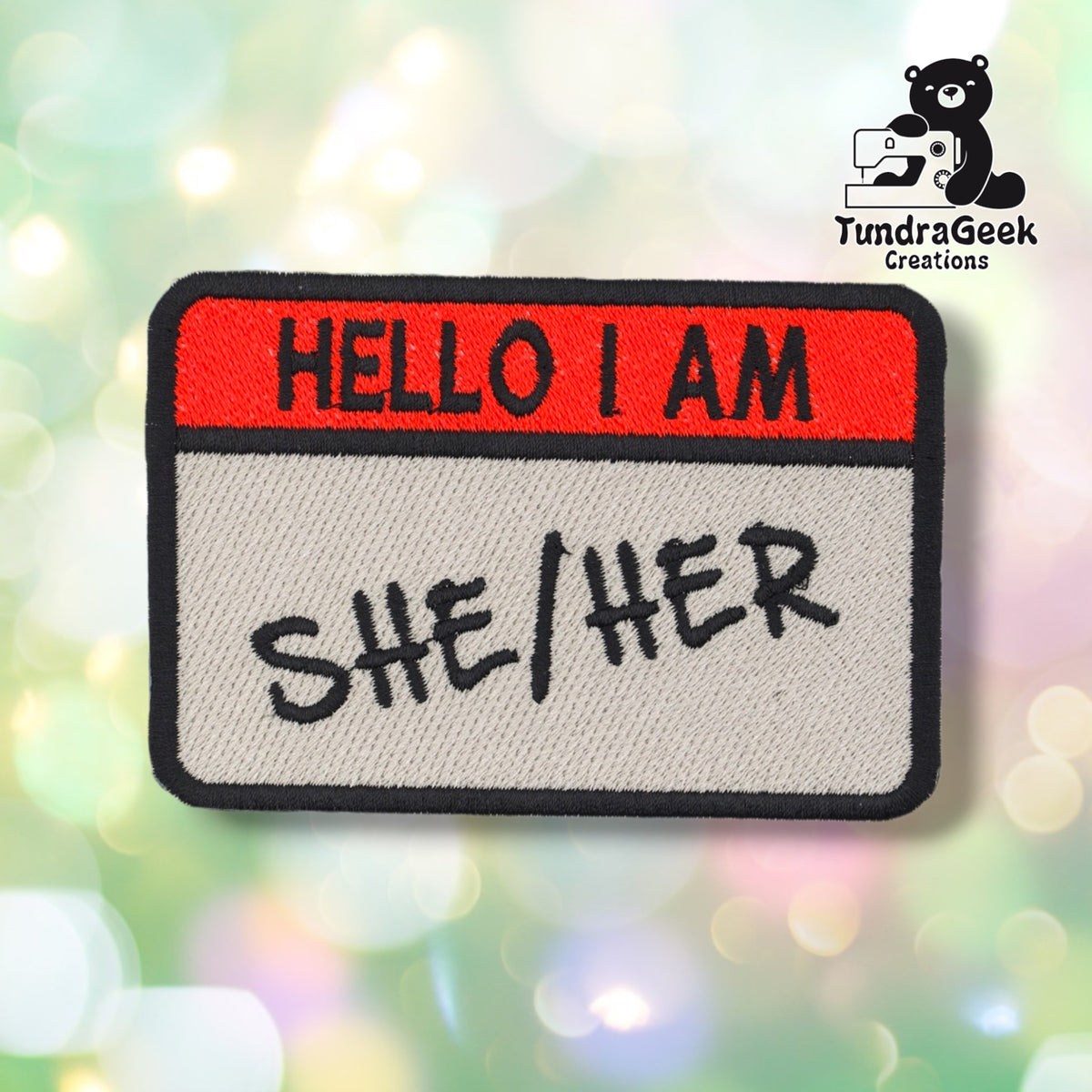 Front view with "She/Her" embroidered in black beneath the red "HELLO I AM" header.