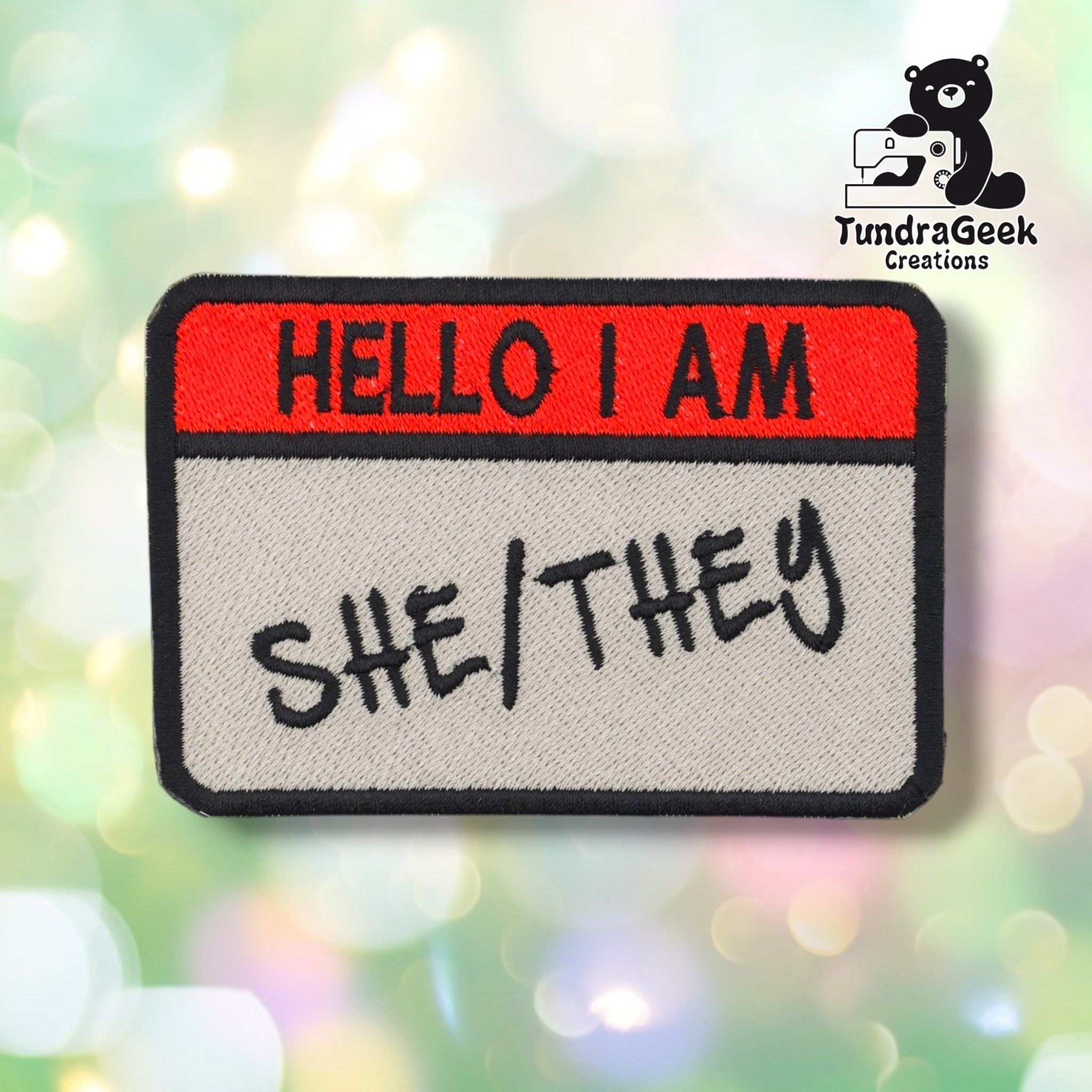 An embroidered patch shaped like a name tag with a red header reading 'HELLO I AM' and a light gray body reading 'SHE/THEY' in black script, shown on a colorful blurred bokeh background. A TundraGeek Creations logo appears in the top right.