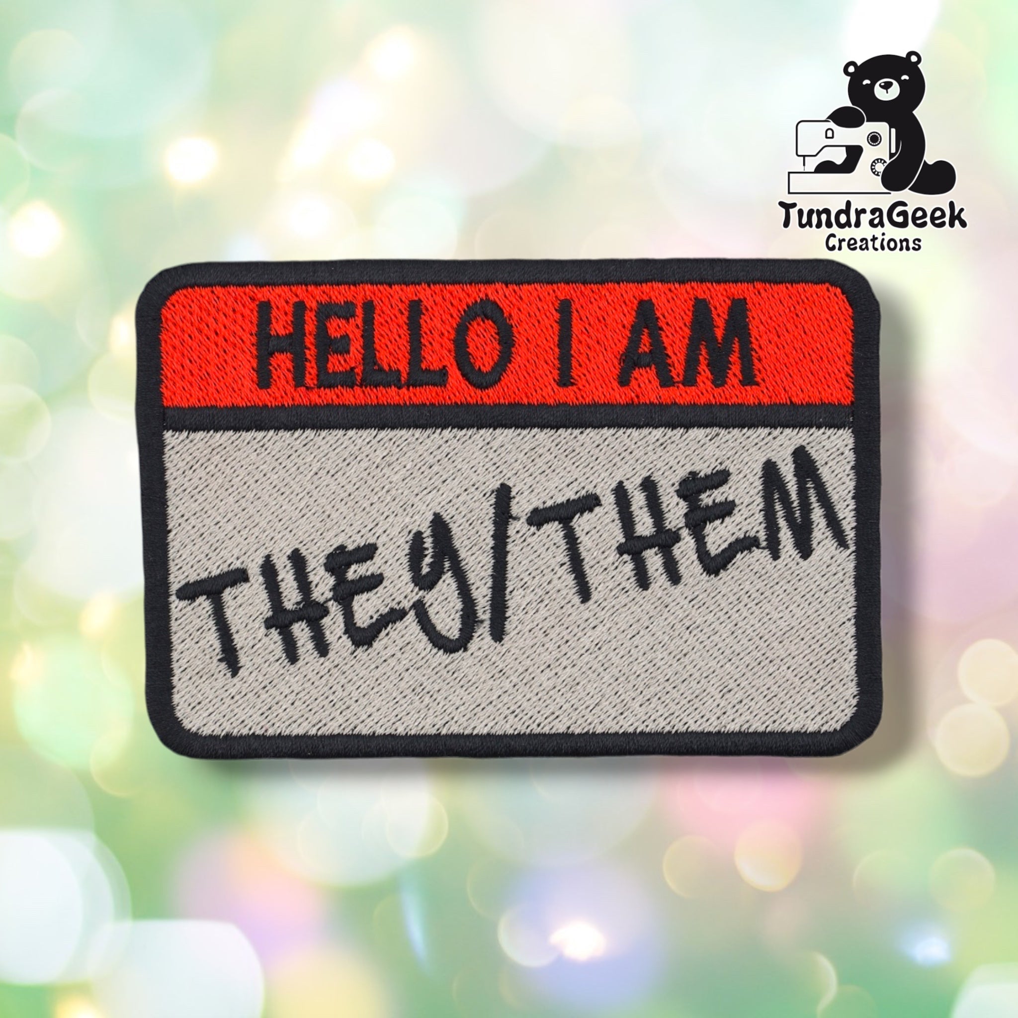 An embroidered patch shaped like a name tag with a red top section reading 'HELLO I AM' and a gray bottom section reading 'THEY/THEM' in black script, shown on a blurred multicolor bokeh background. A TundraGeek Creations logo appears in the top right corner.