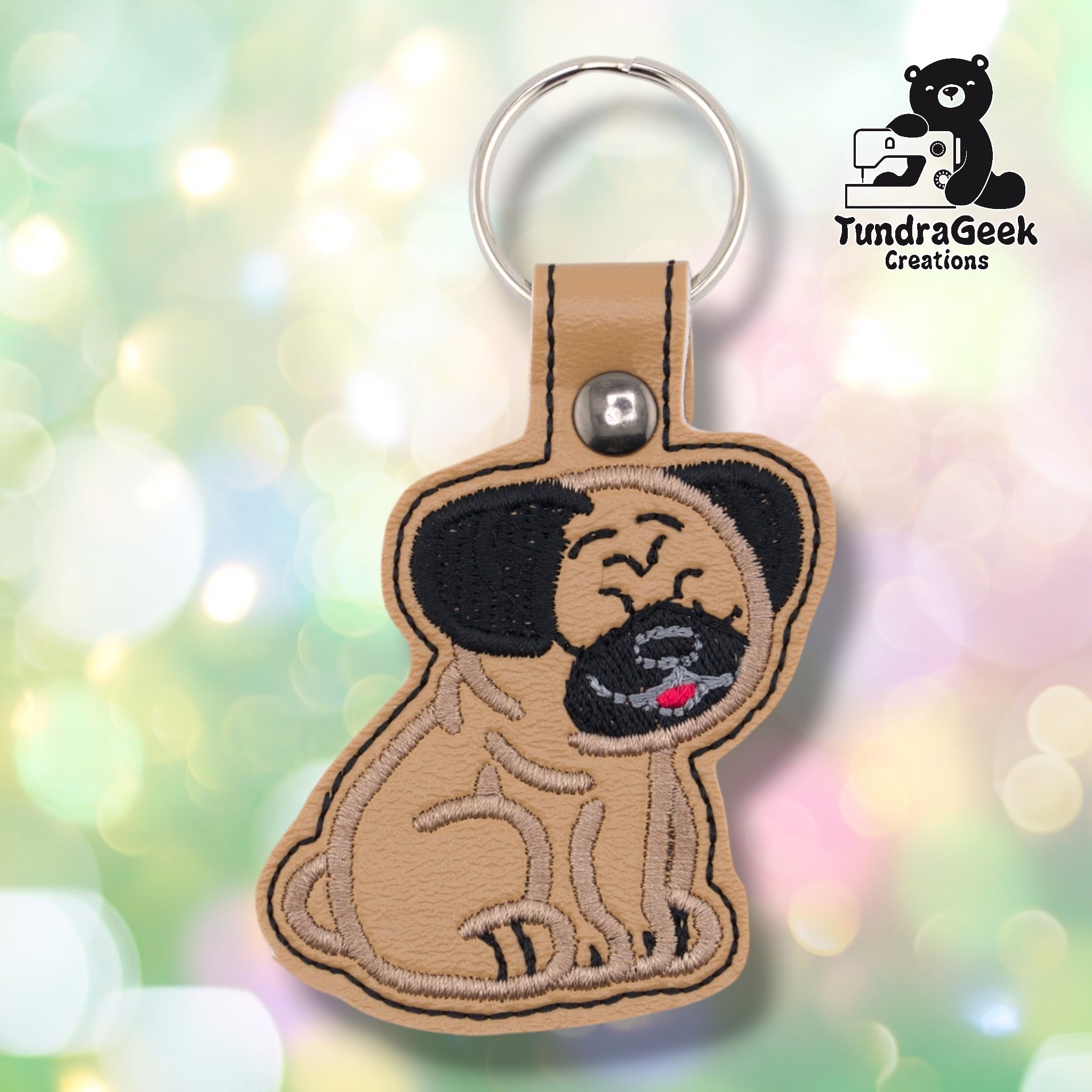 Tan faux leather keychain shaped like a sitting pug. Large black embroidered ears flank a tan wrinkled face with squinting happy eyes, a dark embroidered muzzle with gray detail, and a small red tongue. Silver metal rivet closure and silver key ring at top. TundraGeek Creations logo in upper right corner.