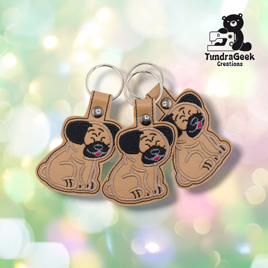 Three pug keychains arranged together on a pastel bokeh background, showing the design from slightly varied angles. All three feature matching black ears, tan base, and silver metal rivet closures.