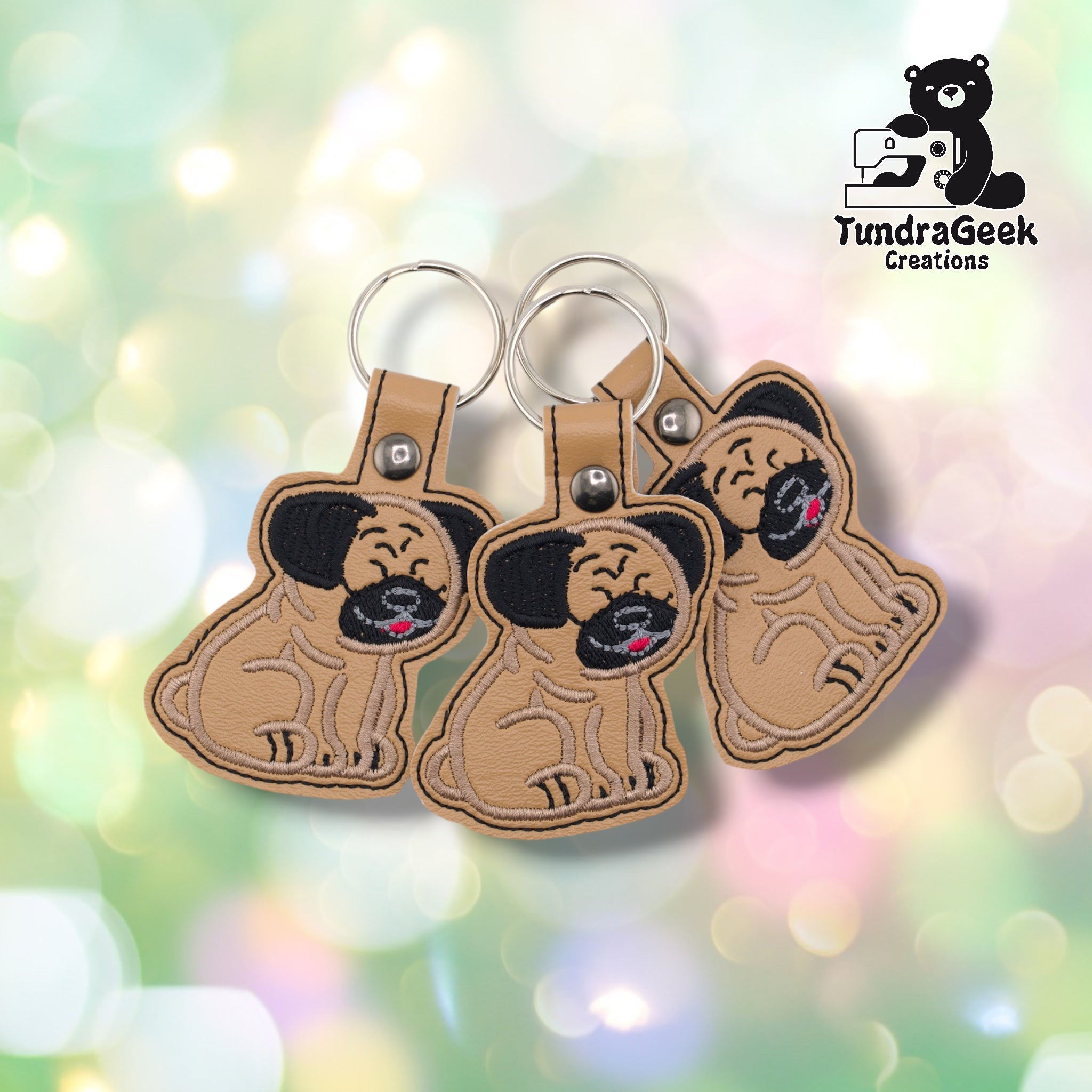 Three pug keychains arranged together on a pastel bokeh background, showing the design from slightly varied angles. All three feature matching black ears, tan base, and silver metal rivet closures.