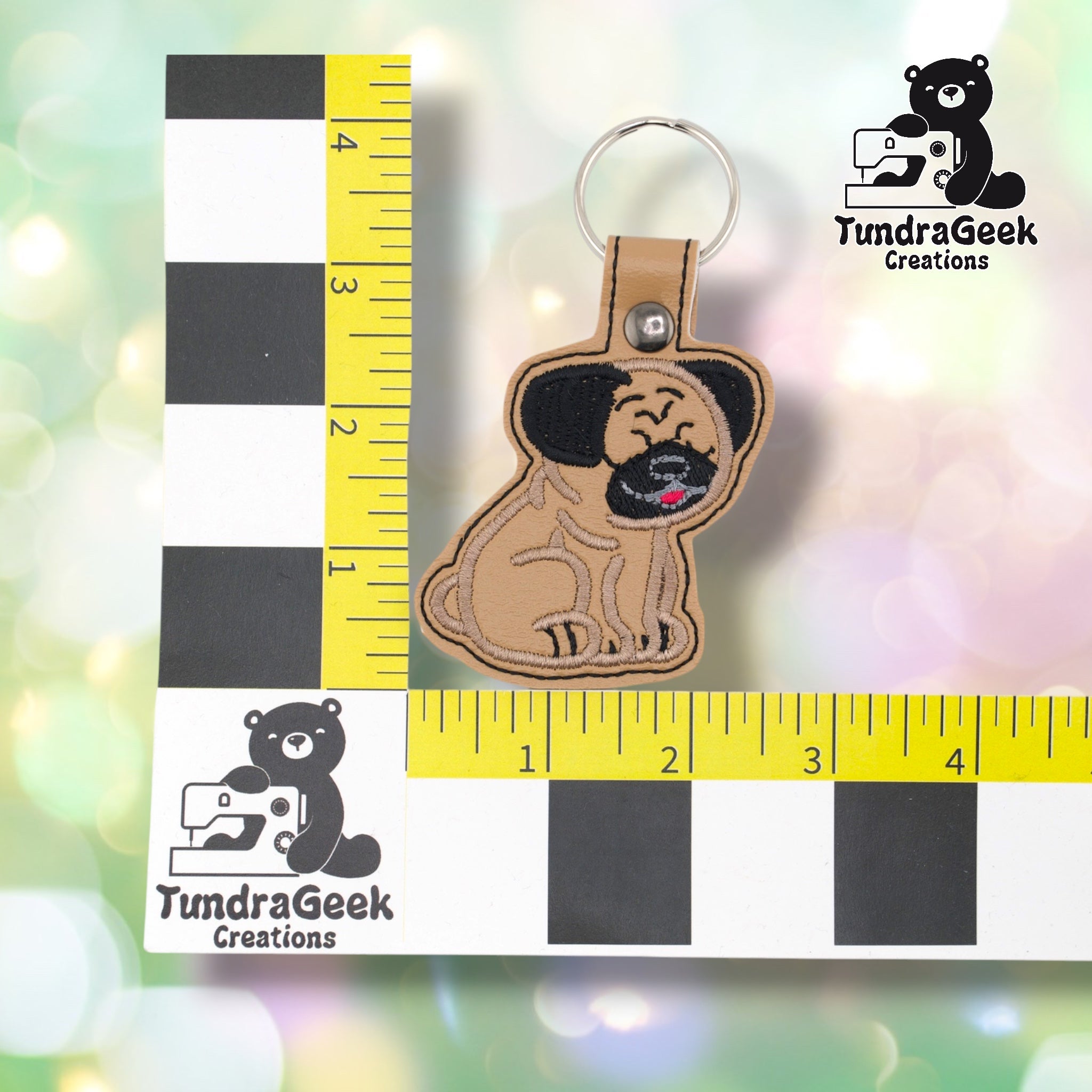 Pug keychain positioned against a TundraGeek Creations branded ruler card showing yellow tape measure. Keychain body measures approximately 2.5 inches wide by 2.5 inches tall, not including hardware.