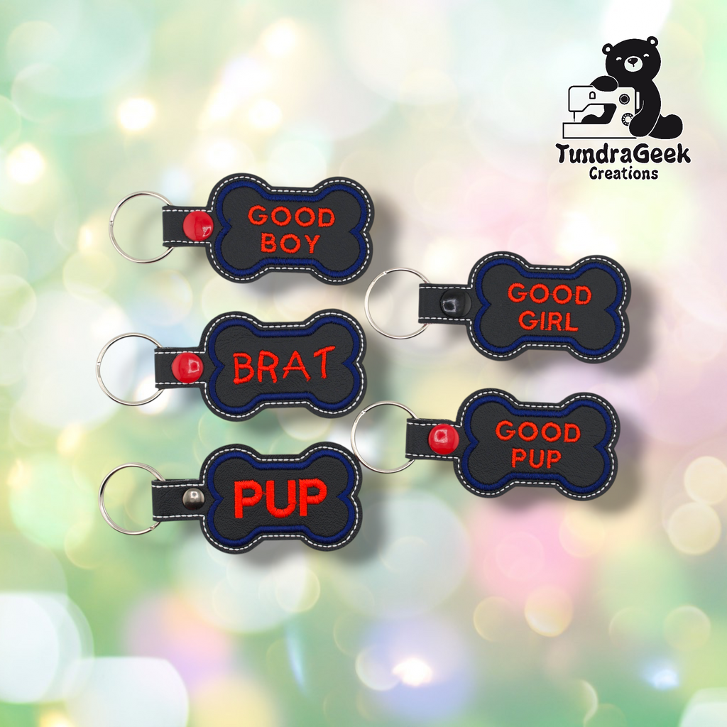 Five pup bone keychains arranged in a loose cluster on a soft bokeh background. From top left: Good Boy (red snap), Good Girl (black snap), Brat (red snap), Pup (silver snap), Good Pup (red snap). All feature black vinyl fronts with blue embroidered borders and red text. Silver split ring hardware on each.