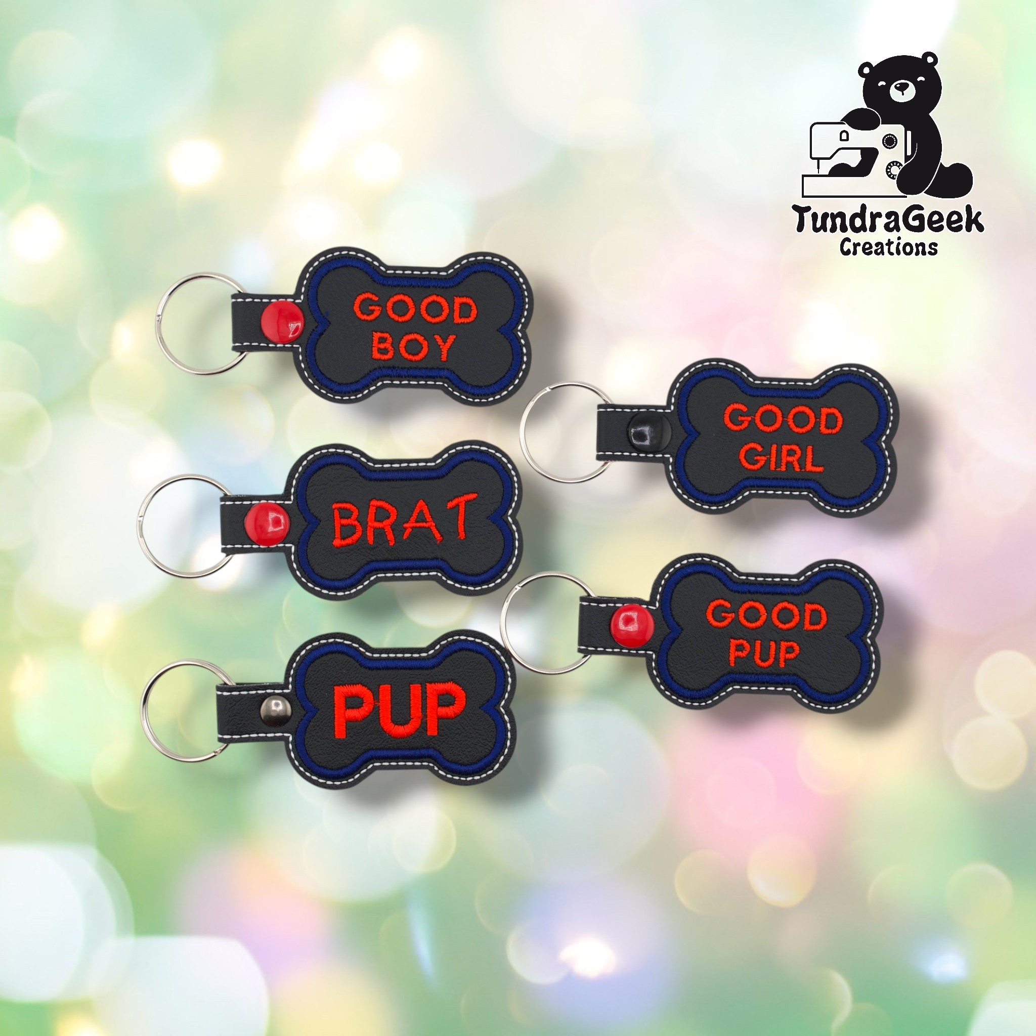 Five pup bone keychains arranged in a loose cluster on a soft bokeh background. From top left: Good Boy (red snap), Good Girl (black snap), Brat (red snap), Pup (silver snap), Good Pup (red snap). All feature black vinyl fronts with blue embroidered borders and red text. Silver split ring hardware on each.
