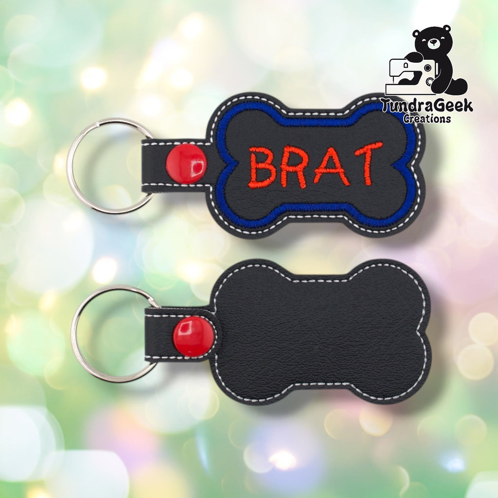 Black vinyl bone-shaped keychain with “BRAT” embroidered in red using a playful hand-lettered style font. Blue embroidered border, white edge stitching. Red snap button and silver split ring hardware on the left.