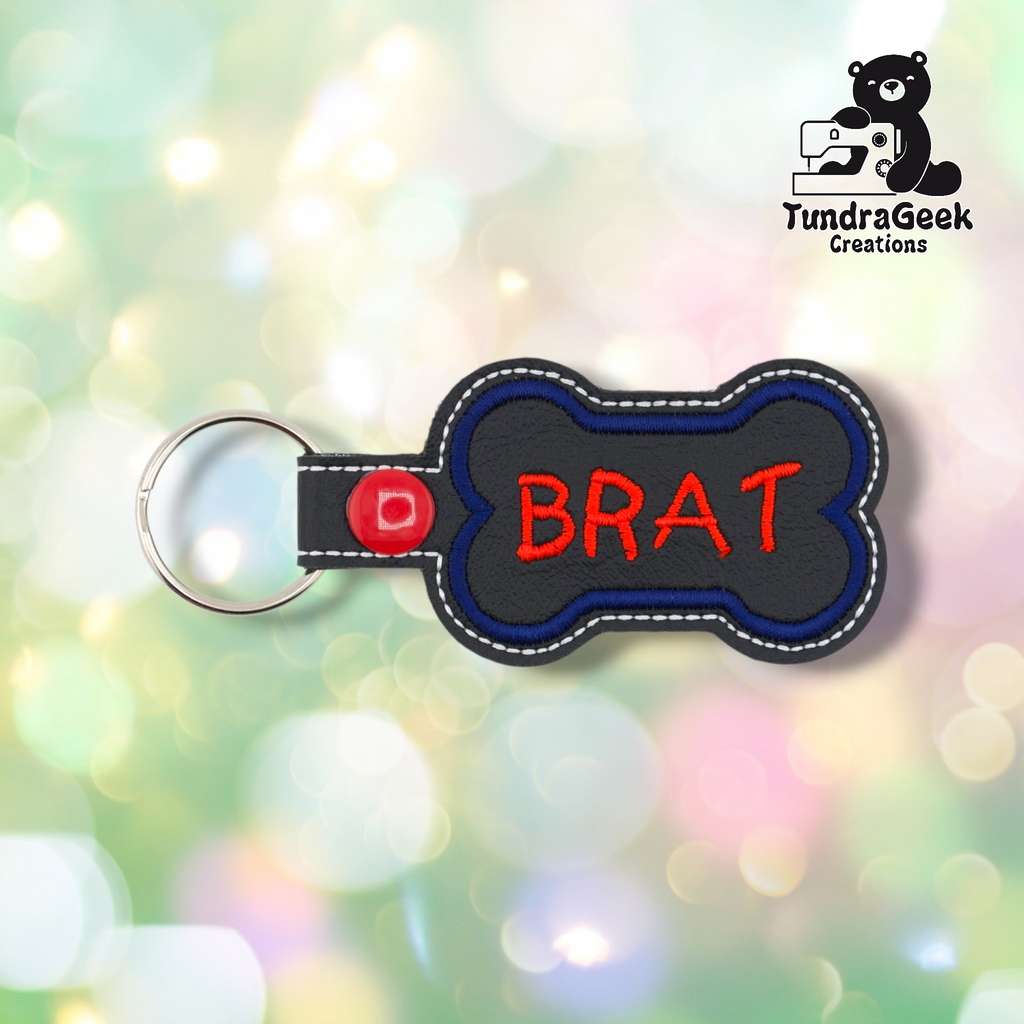 Black vinyl bone-shaped keychain with “BRAT” embroidered in red using a playful hand-lettered style font. Blue embroidered border, white edge stitching. Red snap button and silver split ring hardware on the left.