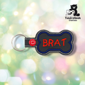 Black vinyl bone-shaped keychain with “BRAT” embroidered in red using a playful hand-lettered style font. Blue embroidered border, white edge stitching. Red snap button and silver split ring hardware on the left.