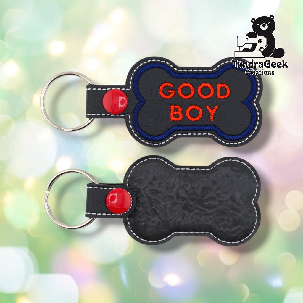 Two views of the “GOOD BOY” keychain. Front: black vinyl with blue border and red “GOOD BOY” text in two lines, white edge stitching, red snap and split ring. Back: black textured vinyl with white edge stitching and red snap hardware.
