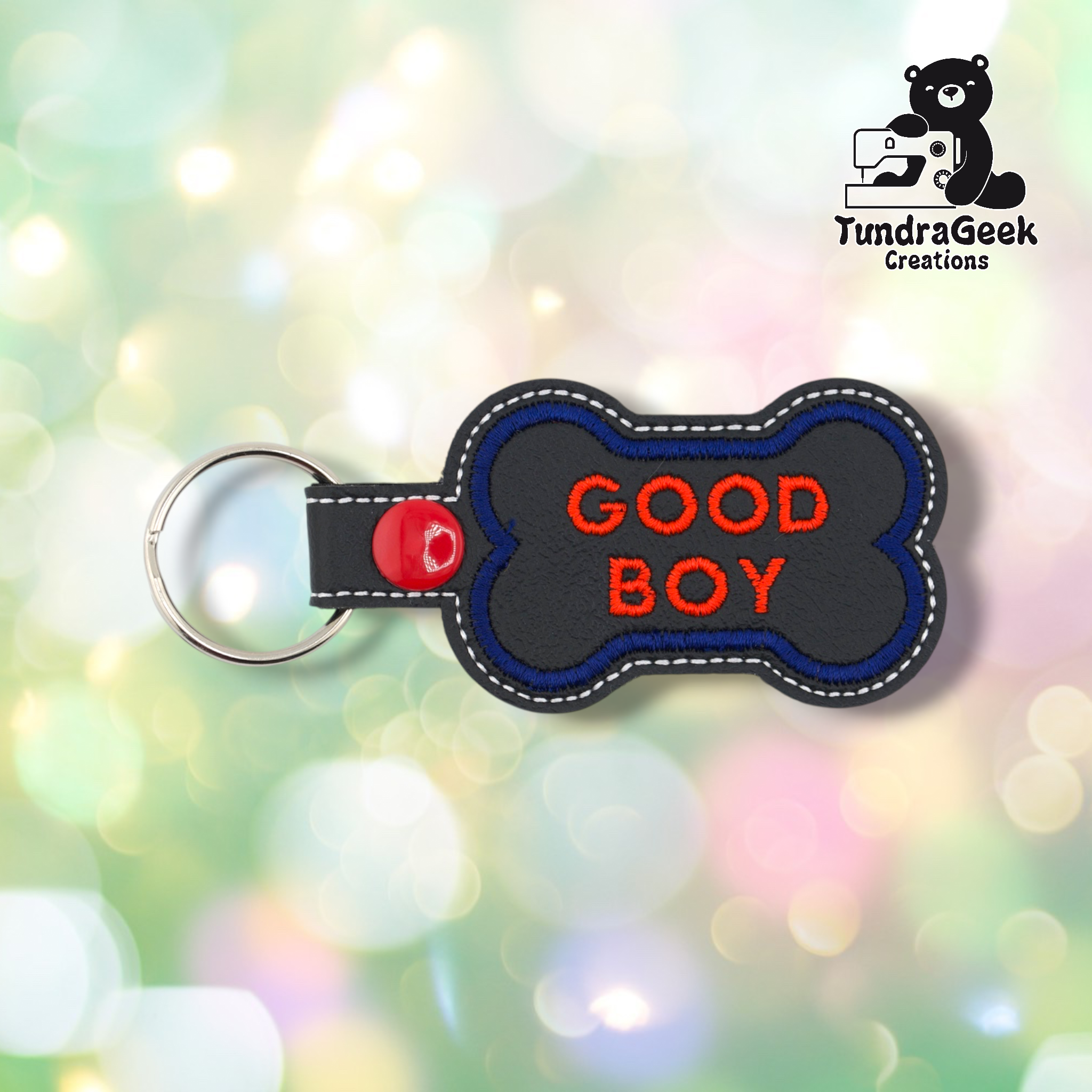 Black vinyl bone-shaped keychain with “GOOD BOY” embroidered in red text in two lines. Blue embroidered border, white edge stitching. Red snap button and silver split ring hardware on the left.