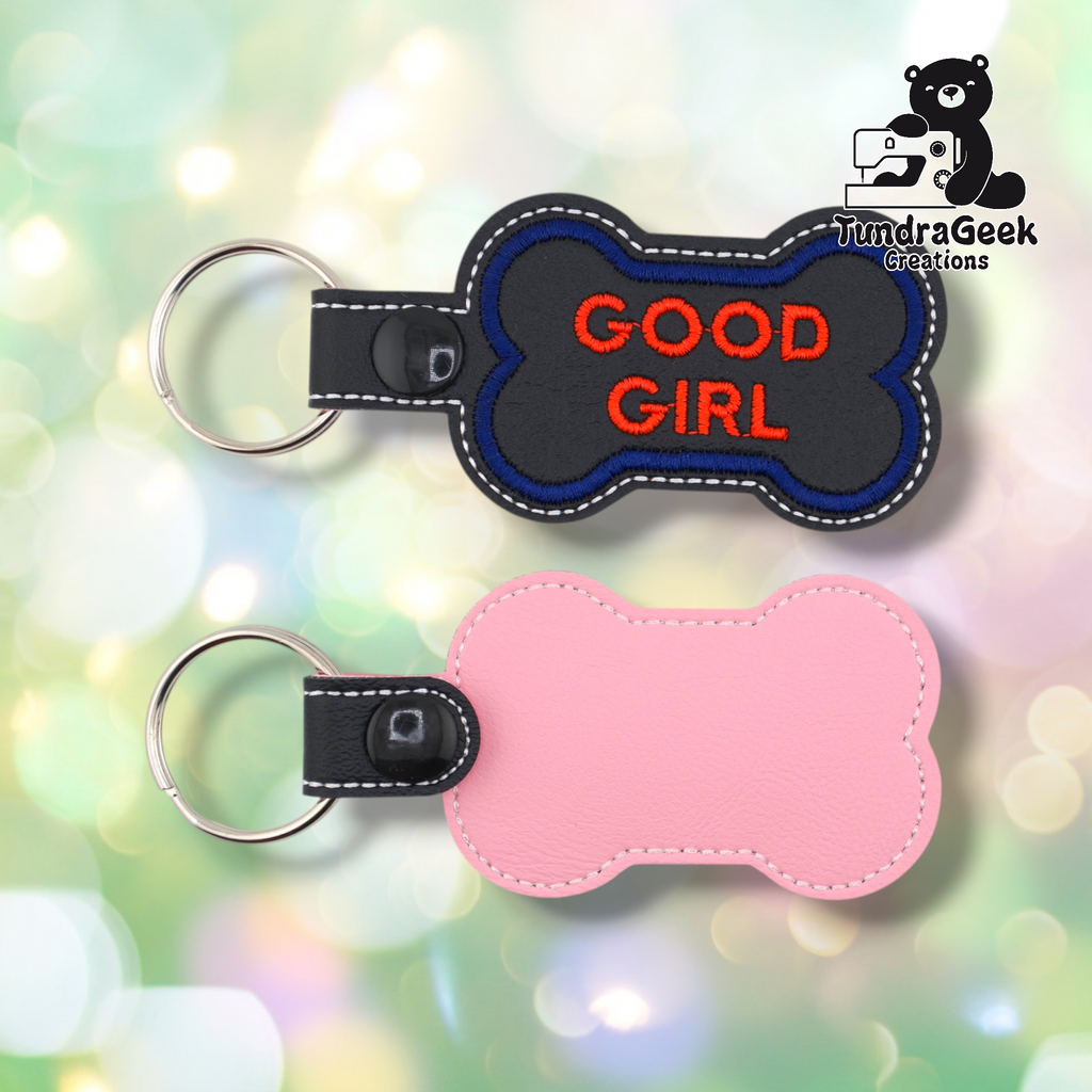 Two views of the “GOOD GIRL” keychain. Front: black vinyl with blue border and red “GOOD GIRL” text in two lines, white edge stitching, black snap and split ring. Back: pink vinyl with white edge stitching and black snap hardware