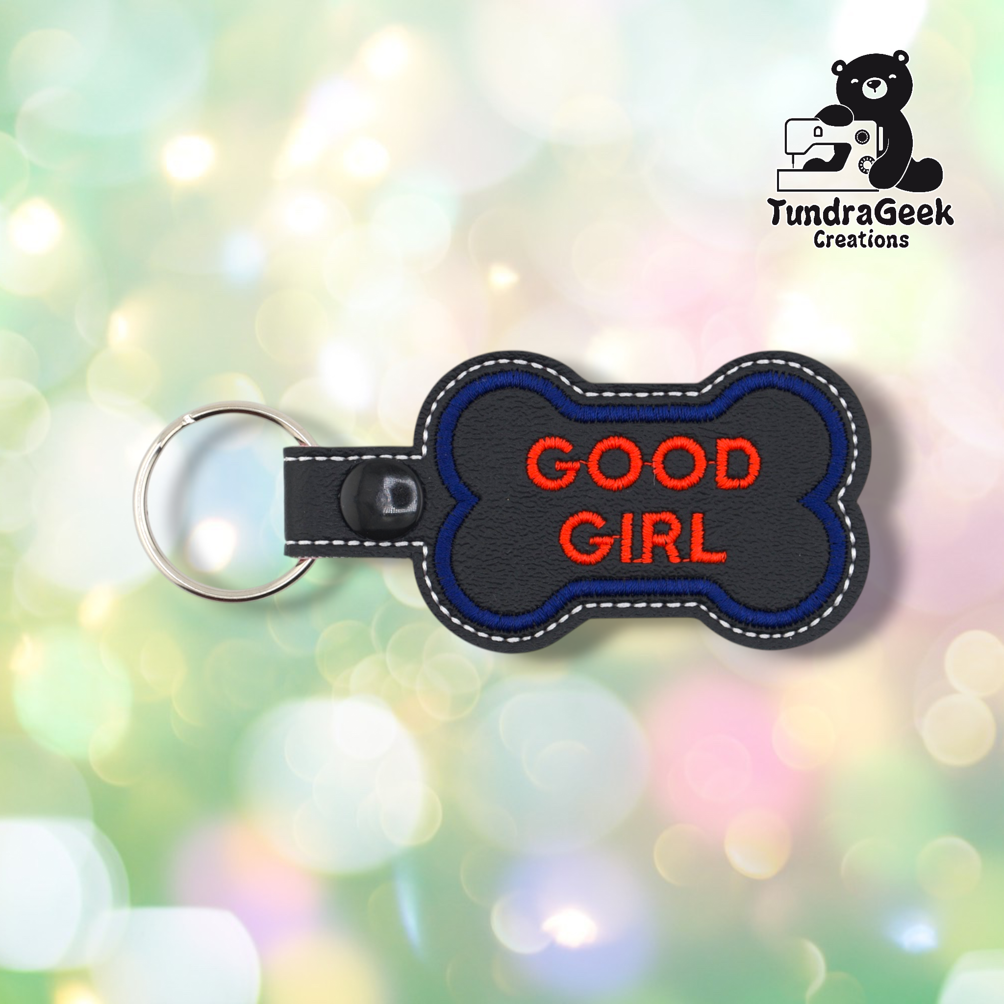 Black vinyl bone-shaped keychain with “GOOD GIRL” embroidered in red text in two lines. Blue embroidered border, white edge stitching. Black snap button and silver split ring hardware on the left.