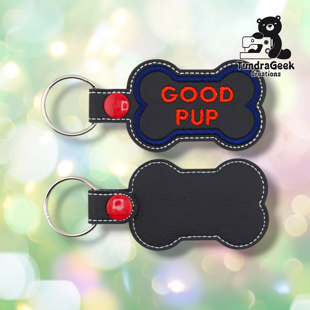 Two views of the “GOOD PUP” keychain. Front: black vinyl with blue border and red “GOOD PUP” text in two lines, white edge stitching, red snap and split ring. Back: plain black vinyl with white edge stitching and red snap hardware