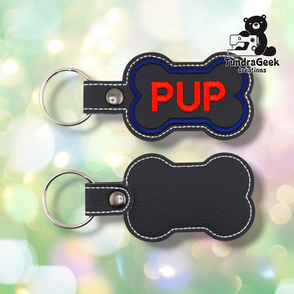 Two views of the “PUP” keychain showing front and back. Front: black vinyl with blue border and red “PUP” text, white edge stitching, silver snap and split ring. Back: plain black vinyl with white edge stitching and silver snap hardware.