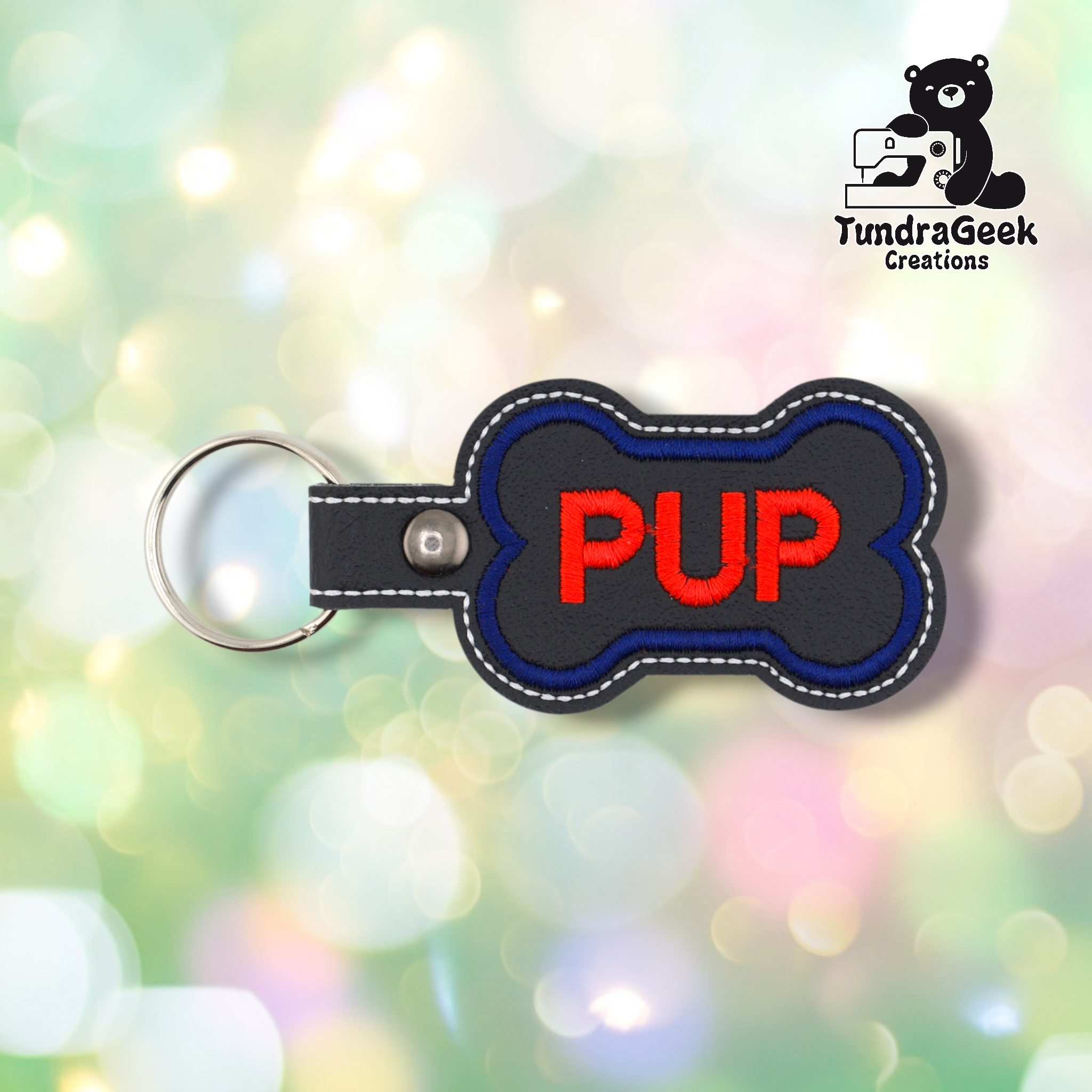Black vinyl dog bone-shaped keychain with “PUP” embroidered in red text on the front. A blue embroidered border follows the bone shape, with white stitching around the outer edge. Silver snap hardware and split ring attached on the left side.