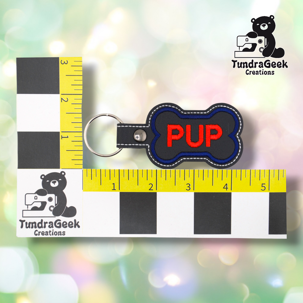 The “PUP” bone keychain shown against a black-and-white checkered background with yellow rulers for scale. The keychain measures approximately 4.25 inches long and 2 inches wide including hardware. Representative of all variants in this listing