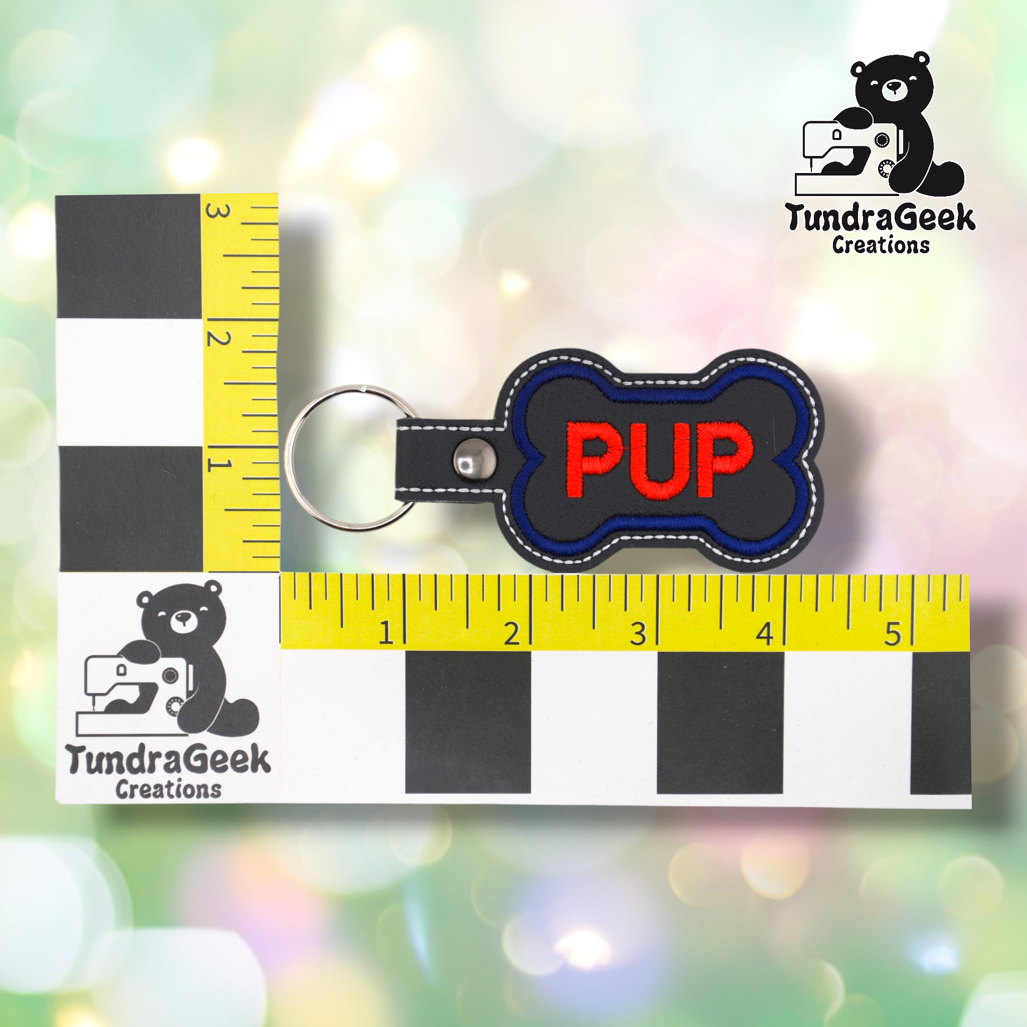 The “PUP” bone keychain shown against a black-and-white checkered background with yellow rulers for scale. The keychain measures approximately 4.25 inches long and 2 inches wide including hardware. Representative of all variants in this listing