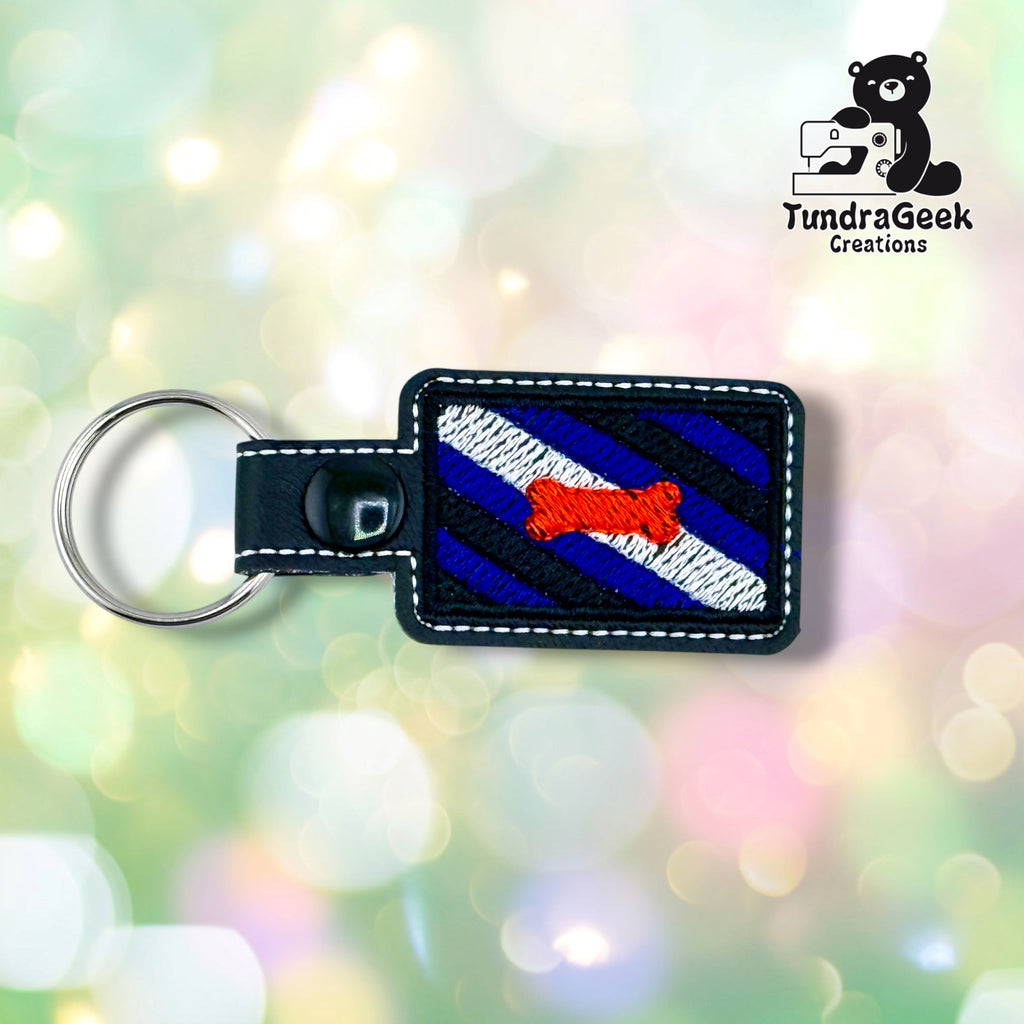 Black rectangular vinyl keychain embroidered with the pup pride flag: diagonal stripes of royal blue and black with a white stripe through the middle and a red-orange dog bone centered on it. White topstitching follows the edge, and a black plastic snap closes the tab around a silver split ring.
