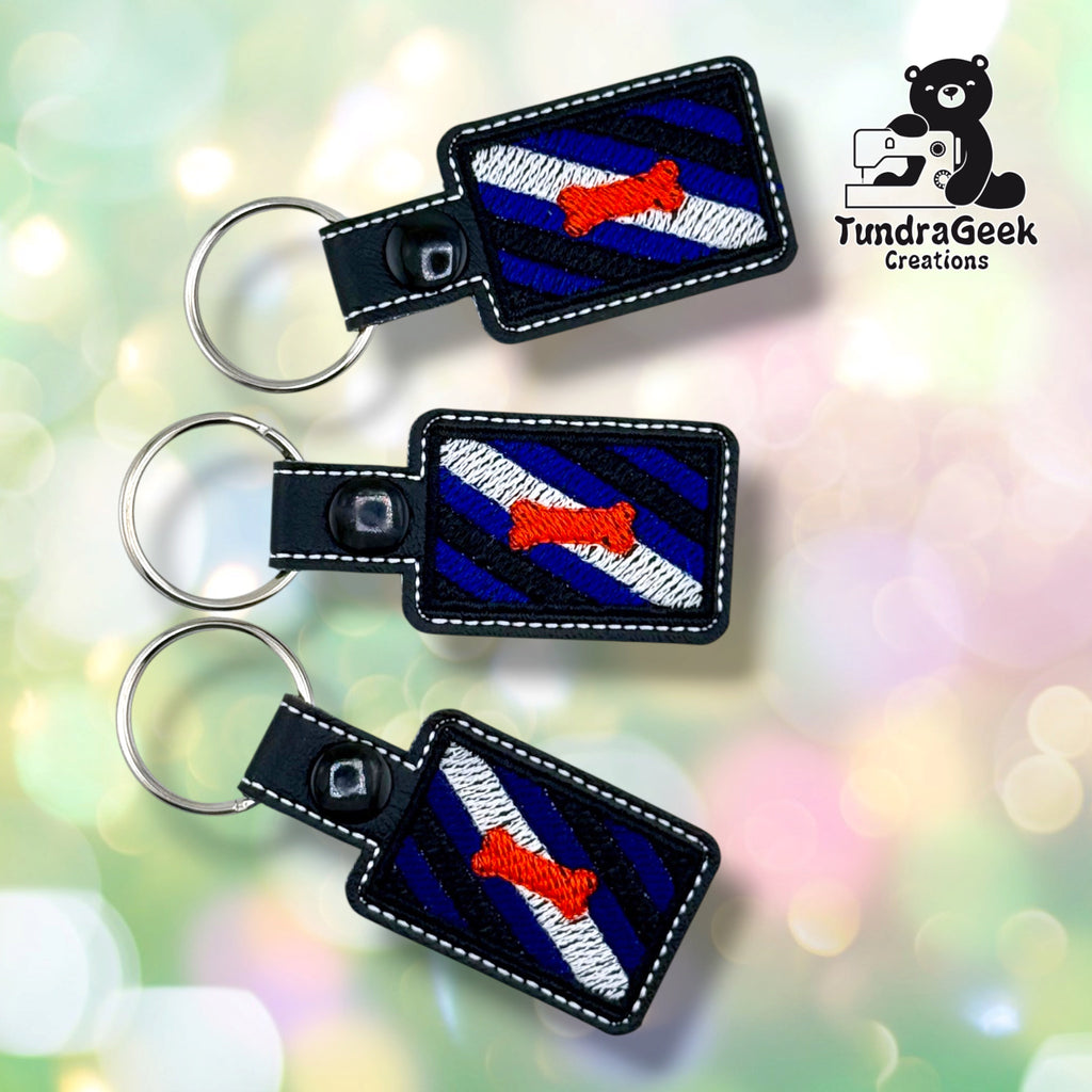Three identical Pup Pride Flag keychains stacked in a column at different angles on a soft bokeh background, each with the same blue, black and white diagonal stripes, red-orange bone, white topstitching and black plastic snap.