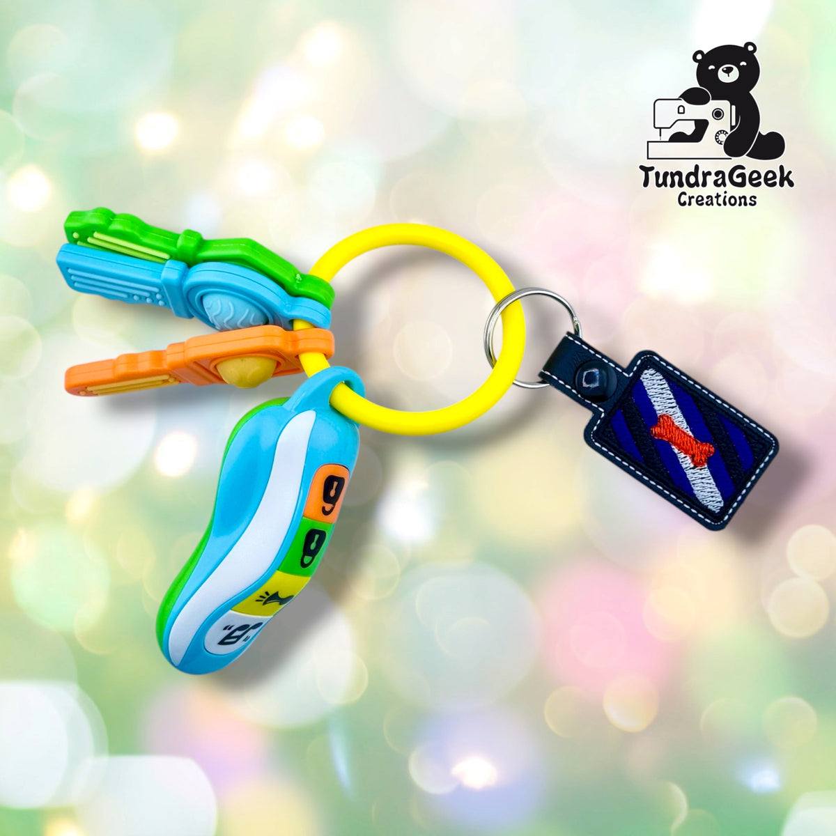 The keychain hanging from a silver split ring on a yellow ring, next to a set of toy plastic keys and a toy key fob in green, blue and orange. The toy keys are a prop for the photo and are not included.