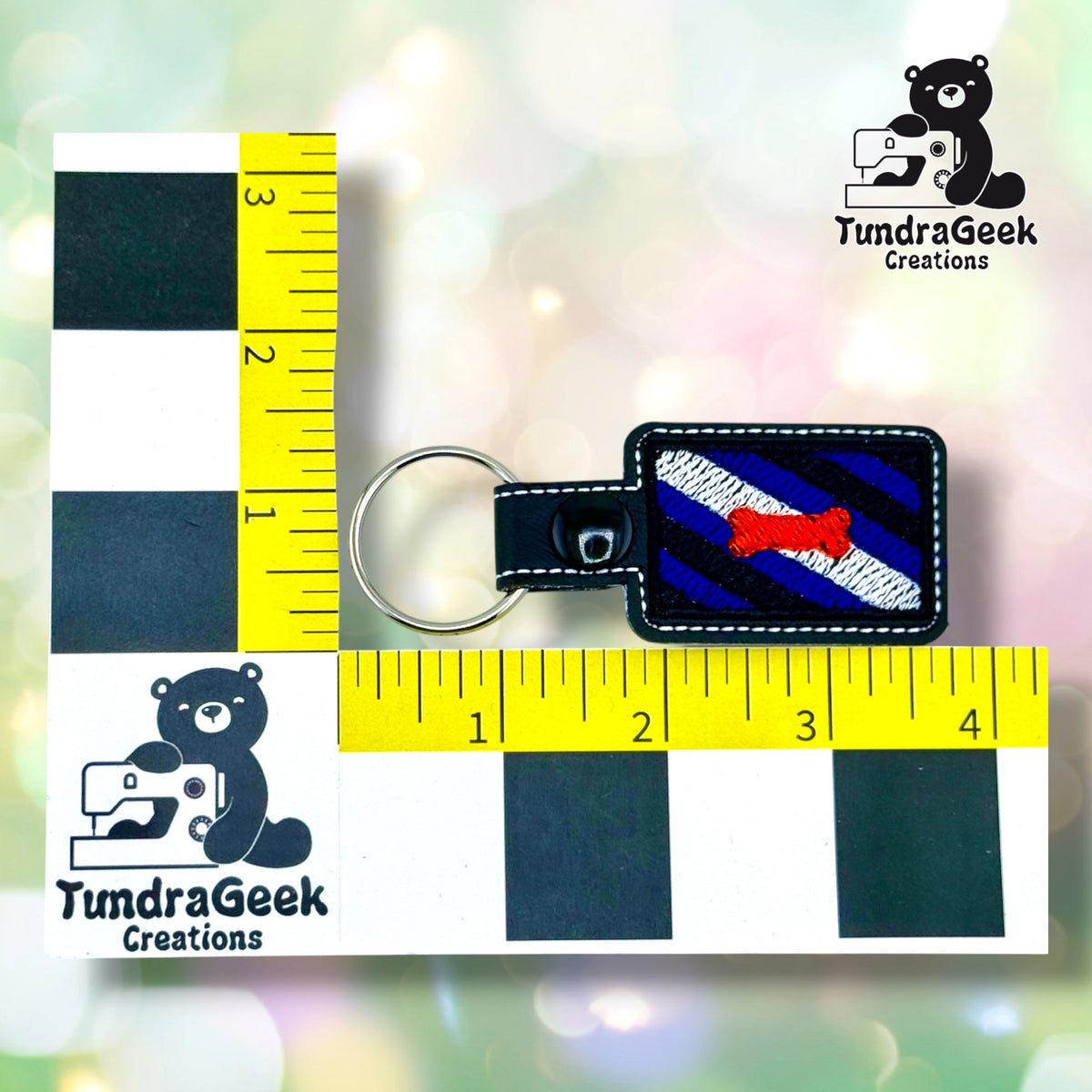 The keychain photographed between two yellow rulers marked in inches, on a black-and-white checkered backing, for size reference. It measures 3.75 inches wide by 1.5 inches tall including hardware.  ---