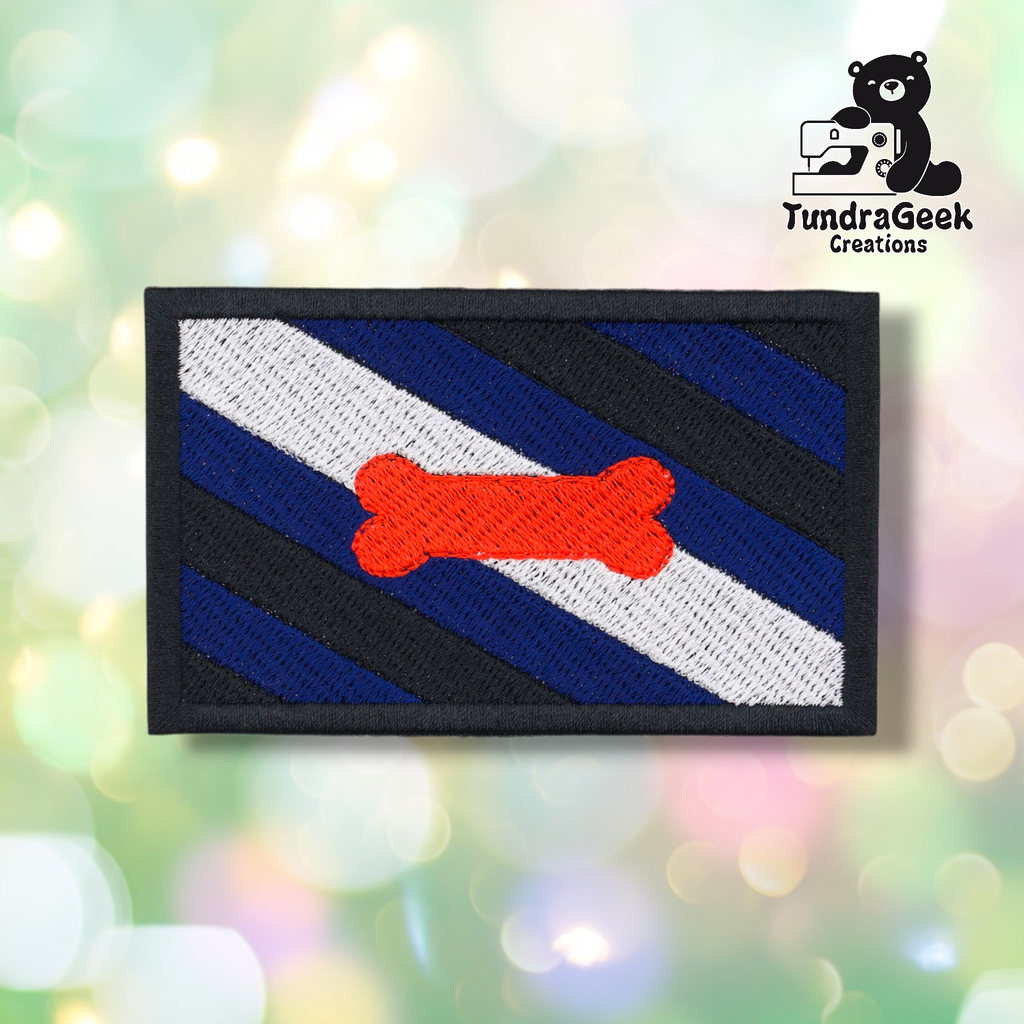 The Pup Pride Flag Patch features diagonal black, blue, and white stripes with an orange bone in the center, plus a TundraGeek Creations cartoon bear logo in the top right. Perfect embroidered sew-on patch for pup play gear with iron-on backing.
