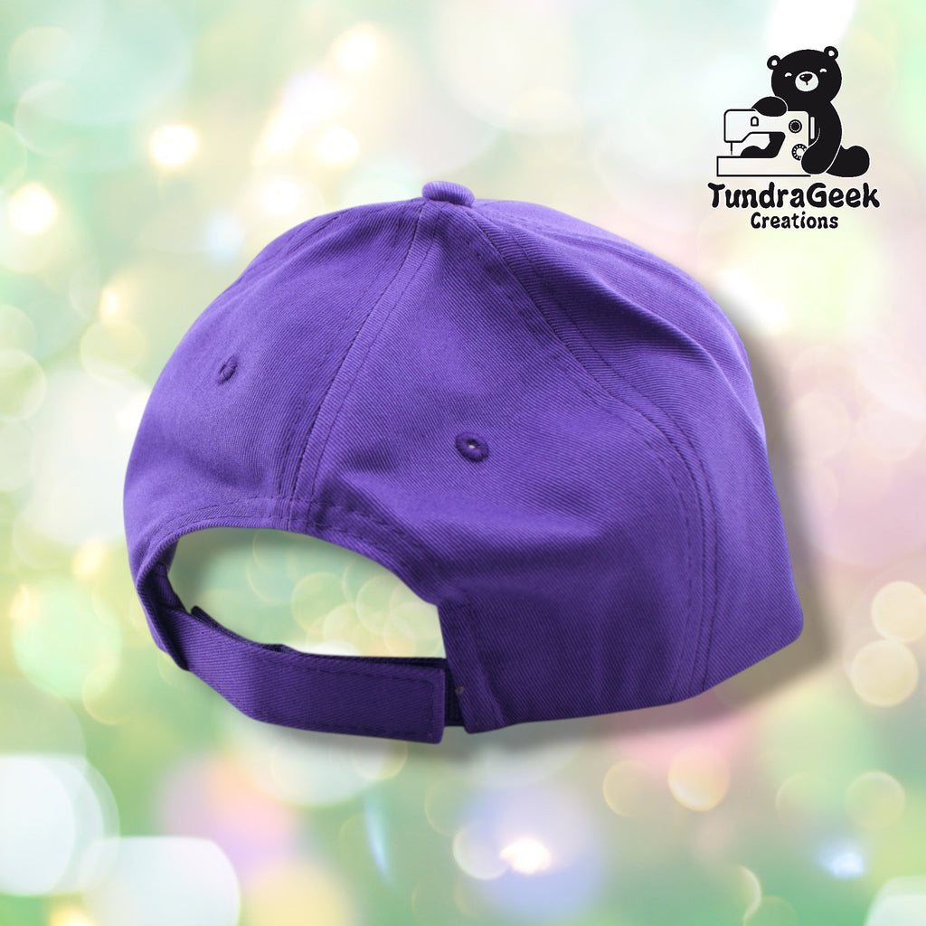A purple  baseball cap displayed against a soft pastel bokeh background with the TundraGeek Creations logo visible in the upper right corner.