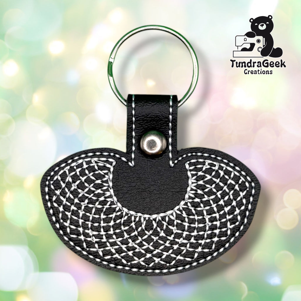 Front of a black vinyl keychain embroidered with a white crocheted-look decorative collar design, scalloped along the bottom edge. Green metal split ring and silver snap closure at top.