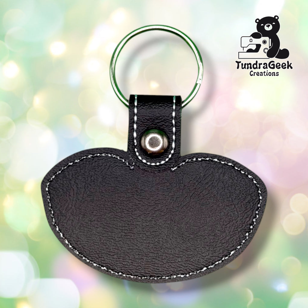 Back of the keychain, showing a plain black vinyl reverse side with white outline stitching following the collar silhouette. Green metal split ring and silver snap closure at top.