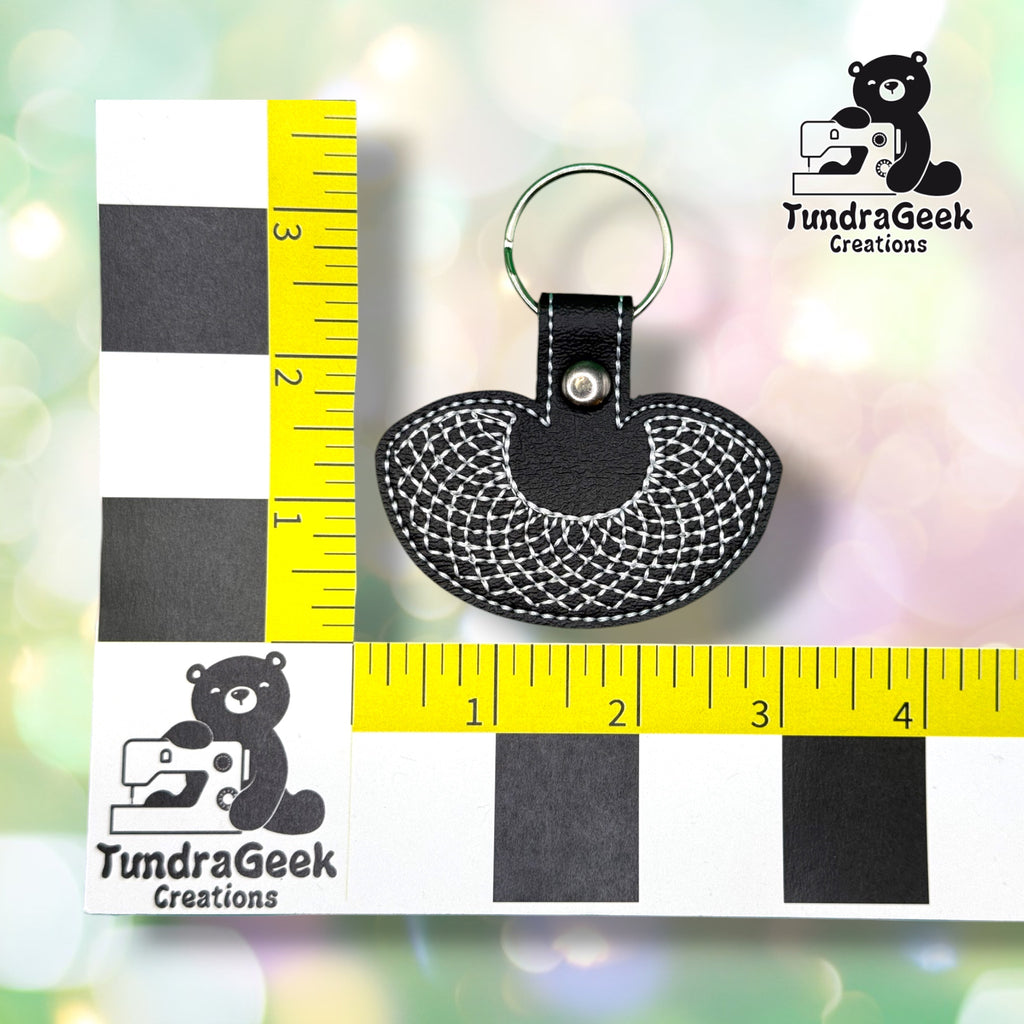 RBG collar keychain shown against a black-and-white checkered background with a yellow ruler for scale, measuring approximately 3.5 inches tall by 3 inches wide.  ---