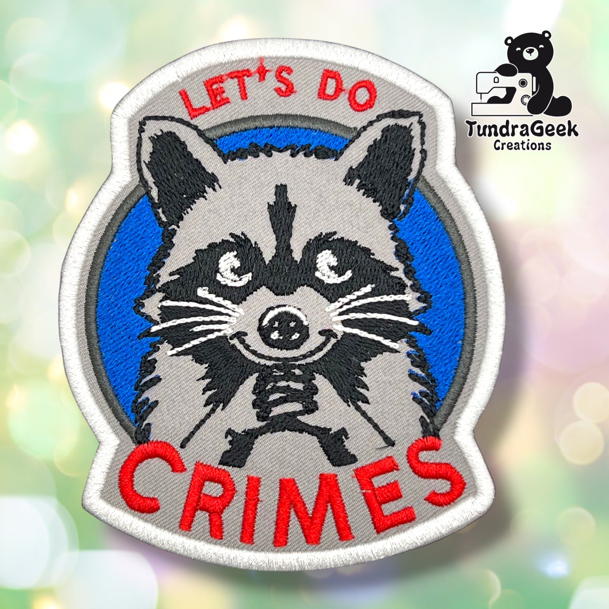 Front of a grey and black patch shaped like a rounded badge, with a blue ring background and a grinning grey raccoon face with black mask markings, white-ringed eyes, and whiskers peeking out from below. "LET'S DO CRIMES" is embroidered in red around the top and bottom.