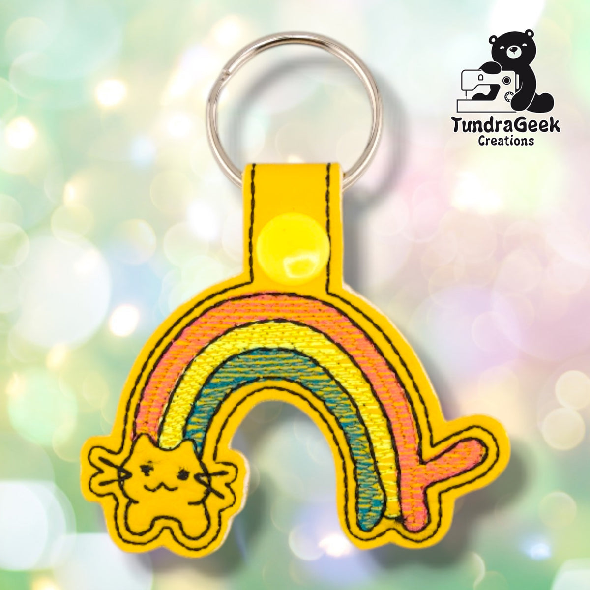 Yellow vinyl keychain embroidered with a pink, yellow, and teal rainbow arcing up from a small cat face with closed eyes and black-outlined whiskers. A yellow plastic snap closes the tab around a silver split ring.