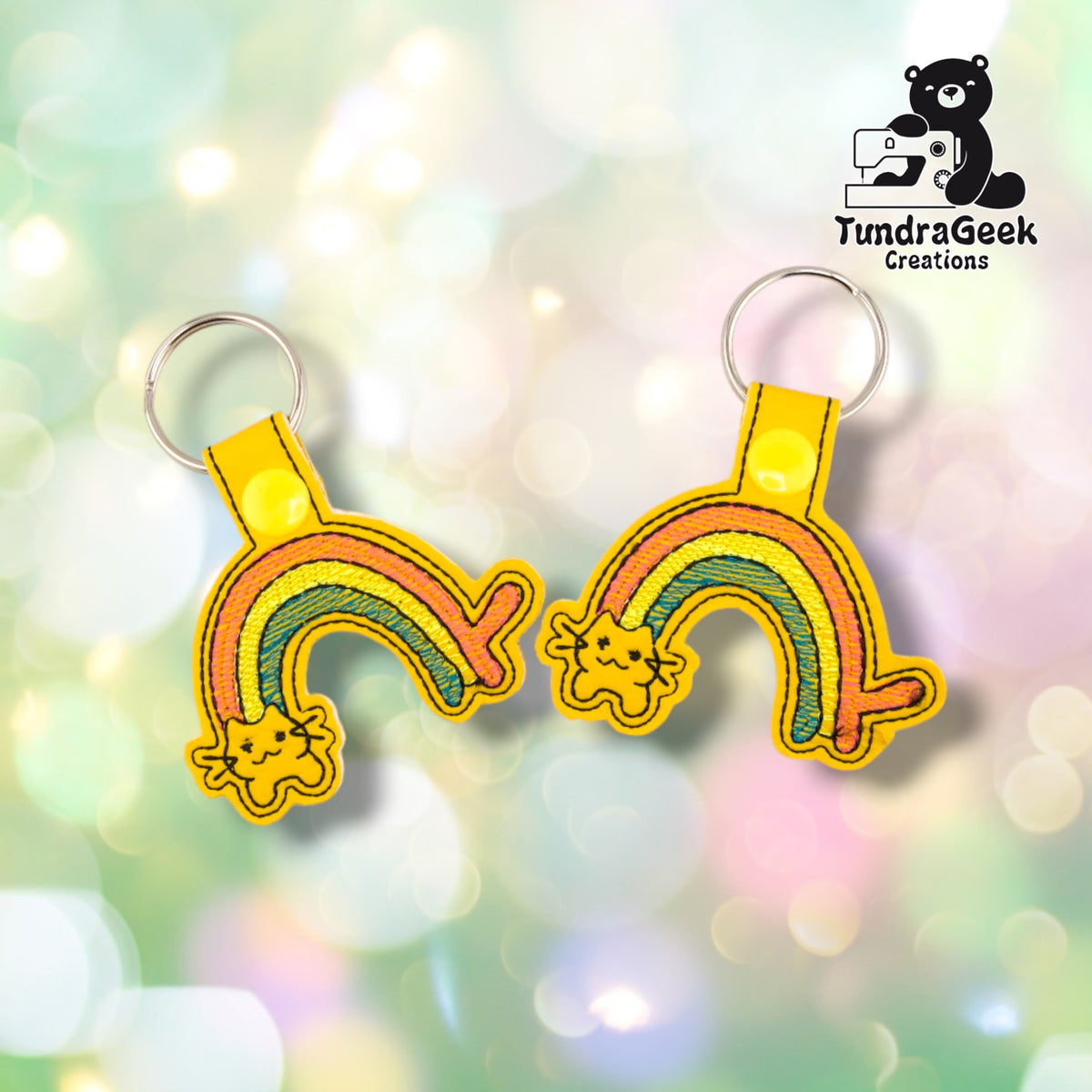 Two Rainbow Kitty keychains shown mirrored, each on yellow vinyl with the same pink, yellow, and teal rainbow and cat face, yellow plastic snap and silver split ring.