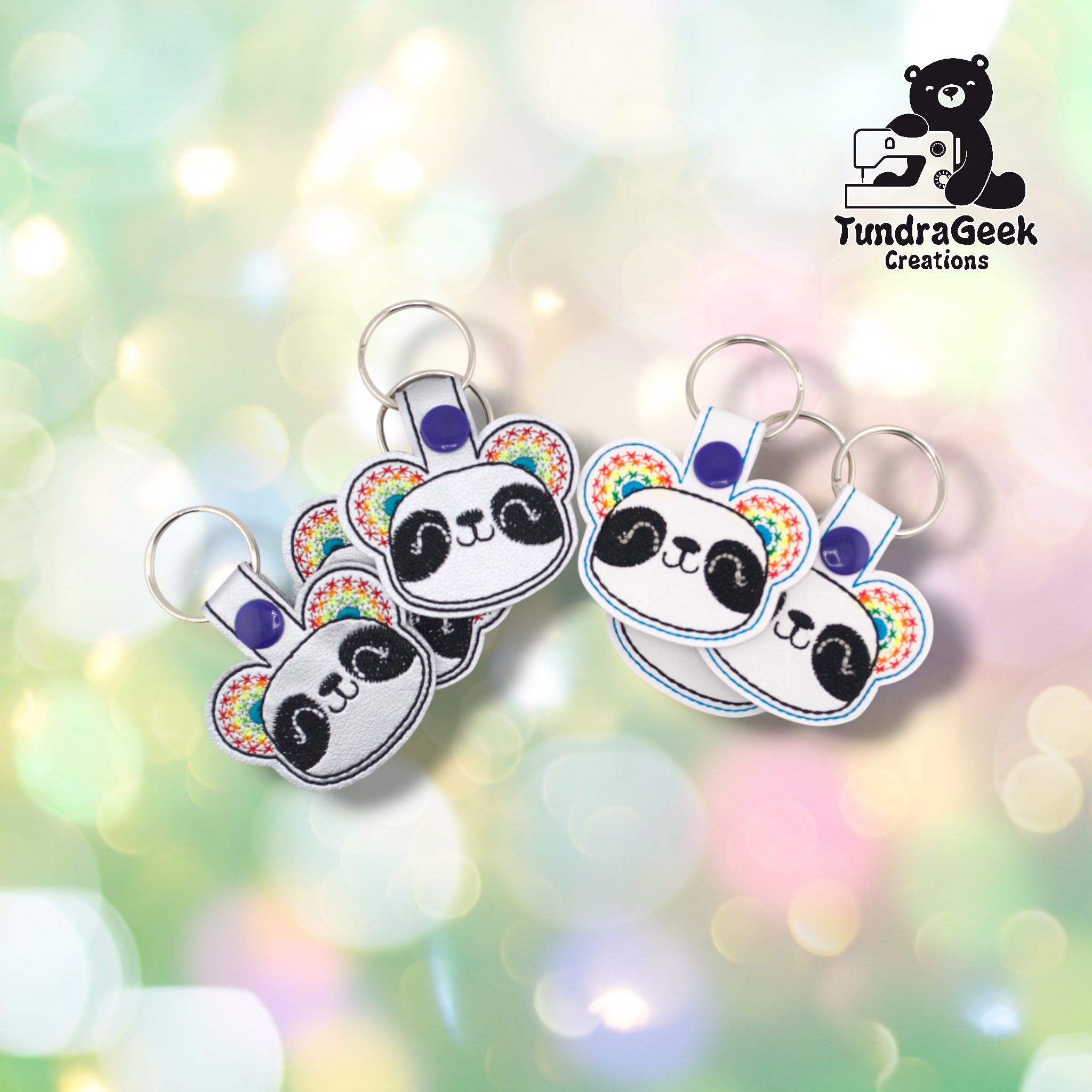 Four rainbow panda keychains arranged in two pairs on a pastel bokeh background. Left pair shows the silver metallic variant; right pair shows the white variant. All feature matching rainbow star-pattern ear embroidery, panda face design, and purple snap closures.