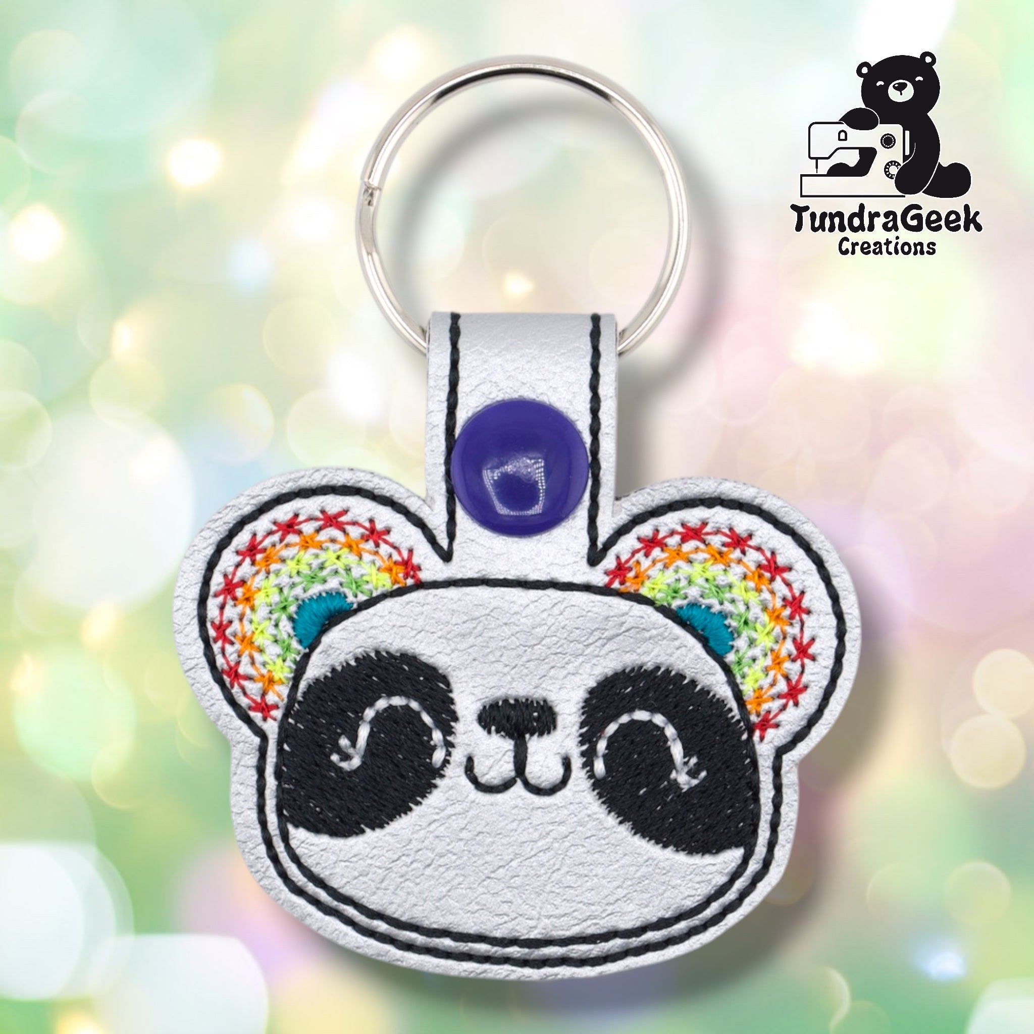 Metallic silver faux leather keychain shaped like a panda face with round ears. Black embroidery details the eye patches, nose, and smile; rainbow star-pattern embroidery with teal accent fills both ears. Purple snap closure and silver key ring at top. TundraGeek Creations logo in upper right corner.