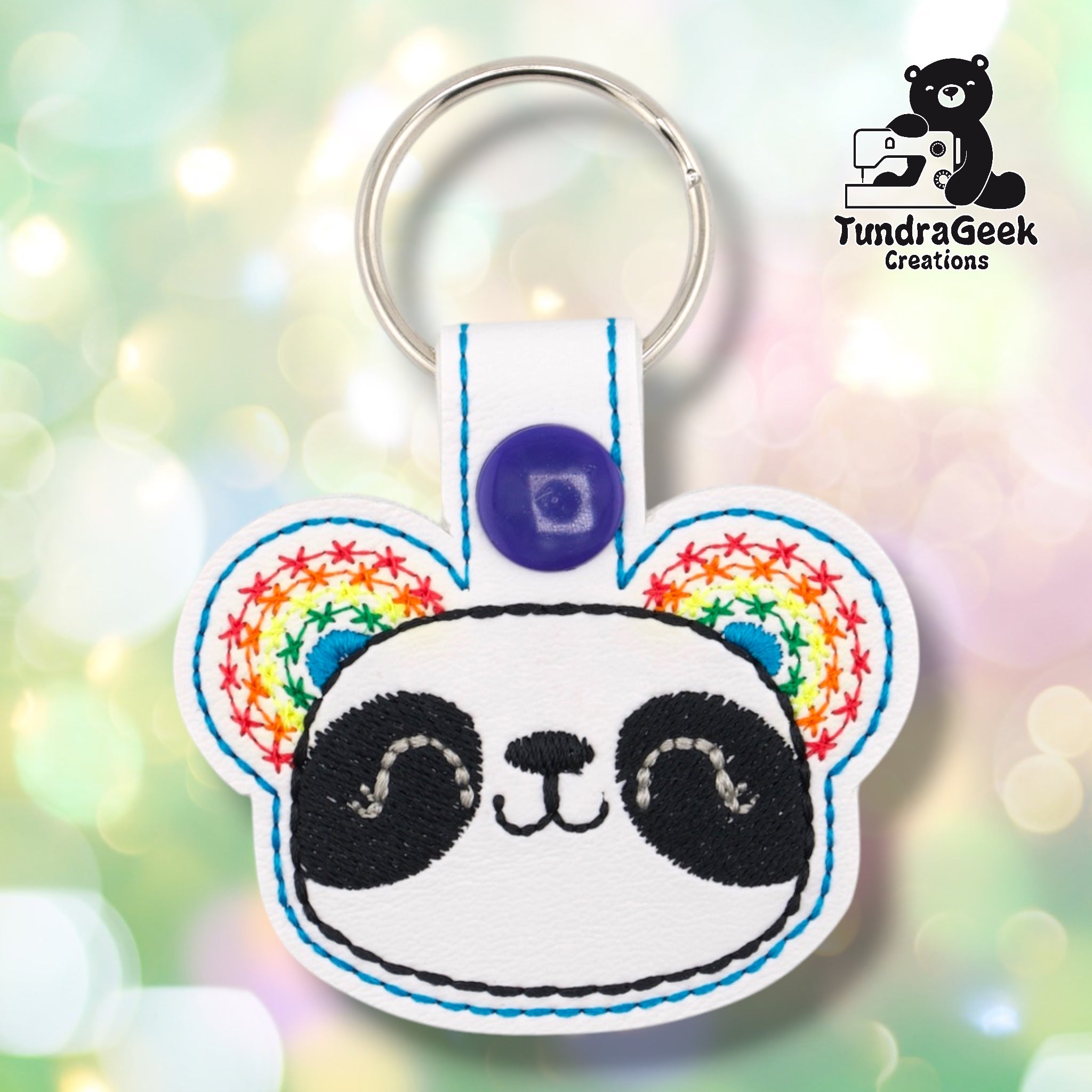 Bright white faux leather keychain shaped like a panda face with round ears. Black embroidery details the eye patches, nose, and smile; rainbow star-pattern embroidery with teal accent fills both ears. Teal outline stitching follows the perimeter. Purple snap closure and silver key ring at top. TundraGeek Creations logo in upper right corner.