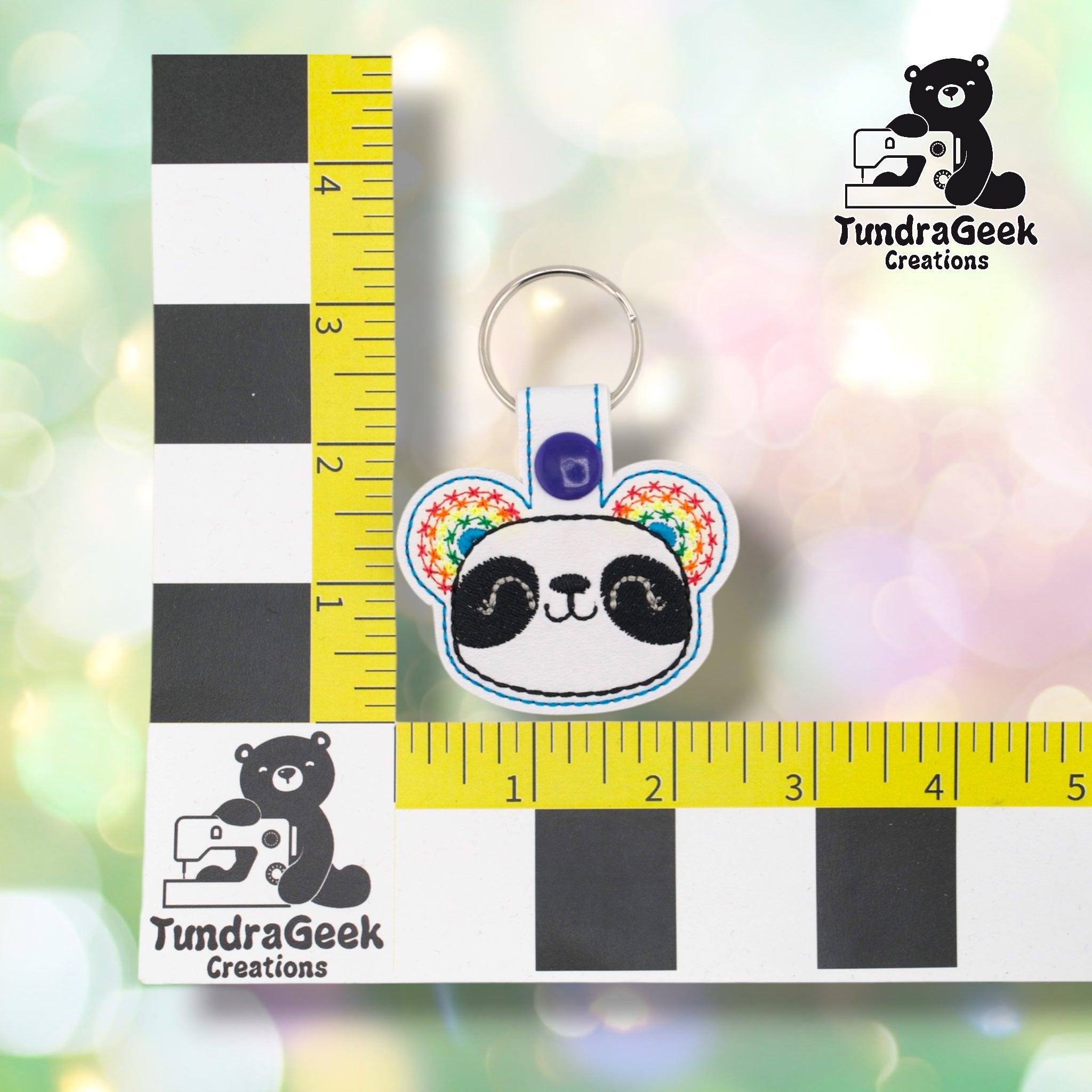 White panda keychain positioned against a TundraGeek Creations branded ruler card showing yellow tape measure. Keychain body measures approximately 2.5 inches wide by 1.75 inches tall, not including hardware.
